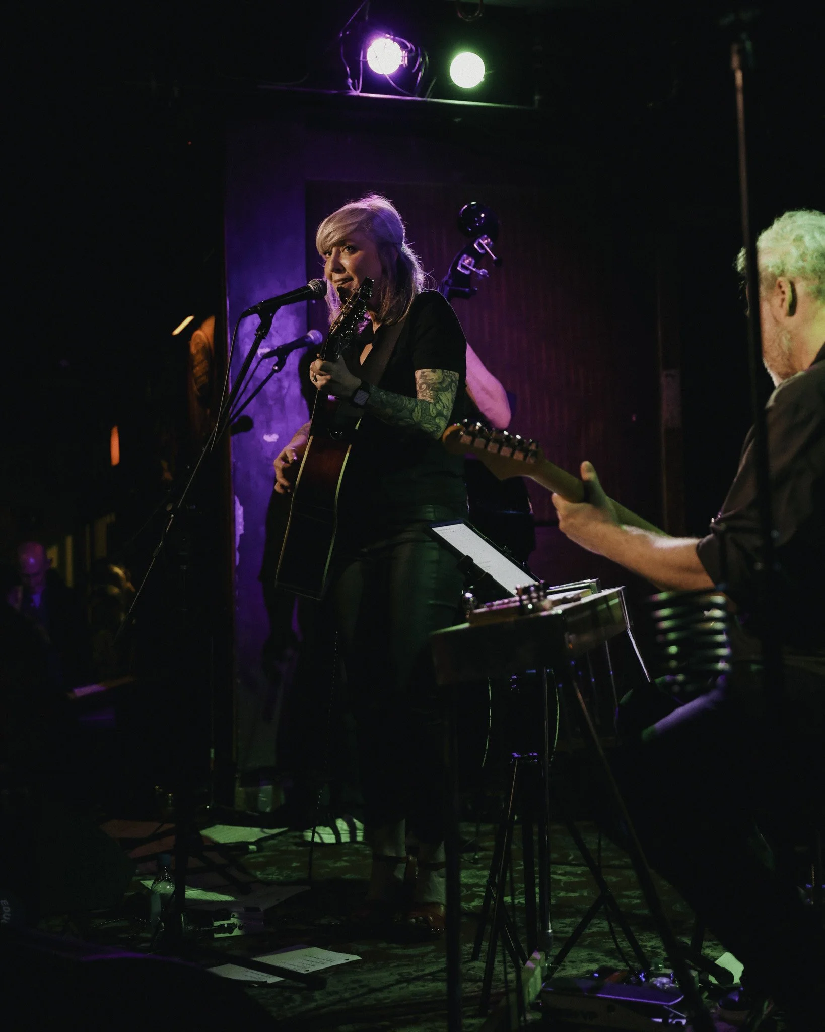 A female musician with tattoos on her arm singing into a microphone while playing an acoustic guitar on stage, with a male musician playing an electric guitar nearby in a dimly lit venue with colorful stage lights.