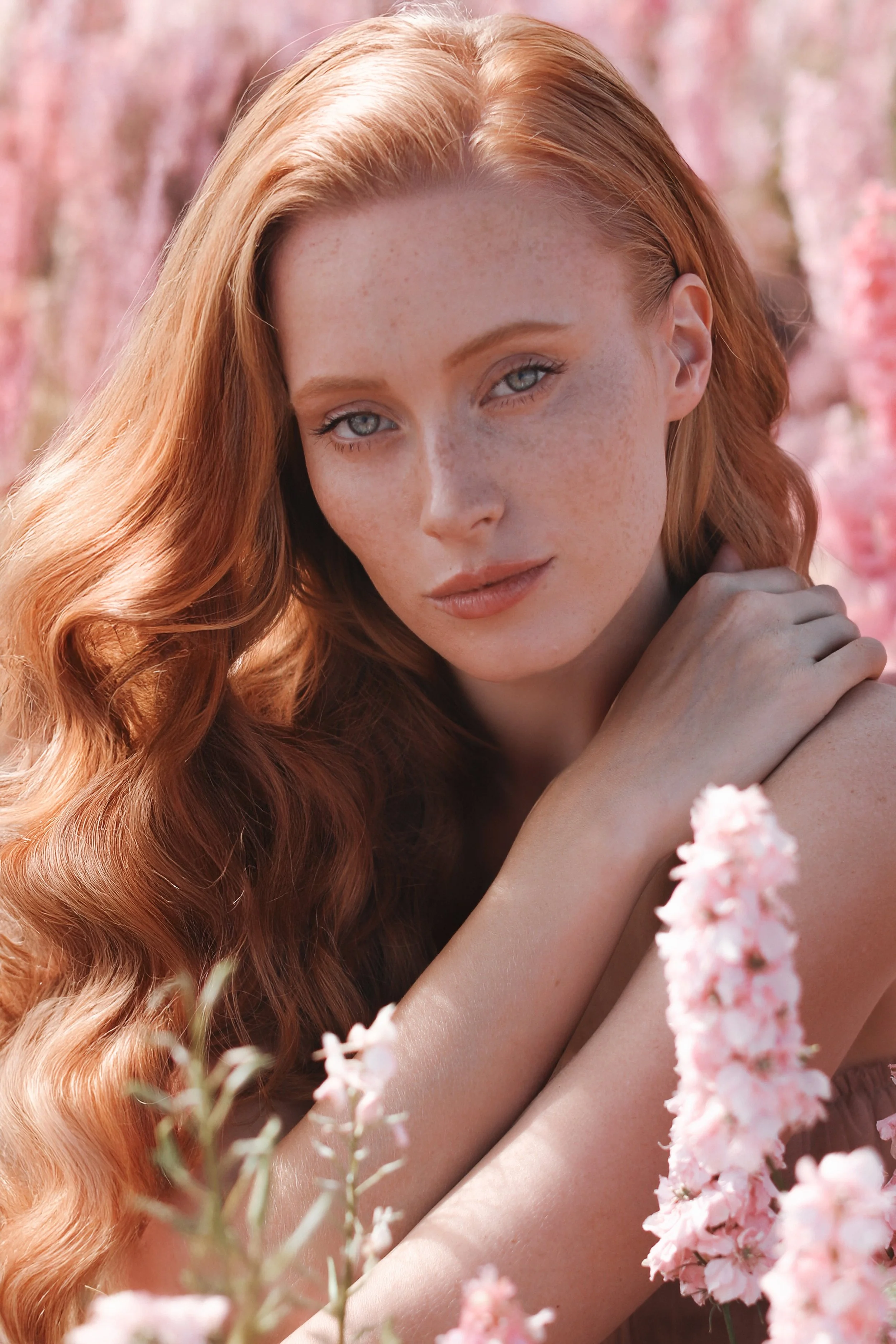 A woman with long, wavy red hair and blue eyes in a field of pink flowers.