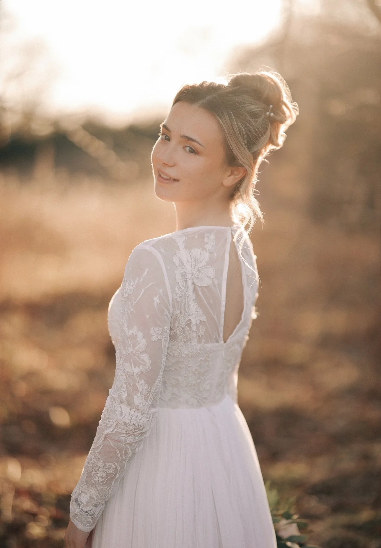 A woman in a white, lace wedding dress standing outdoors during sunset, looking at the camera and smiling.