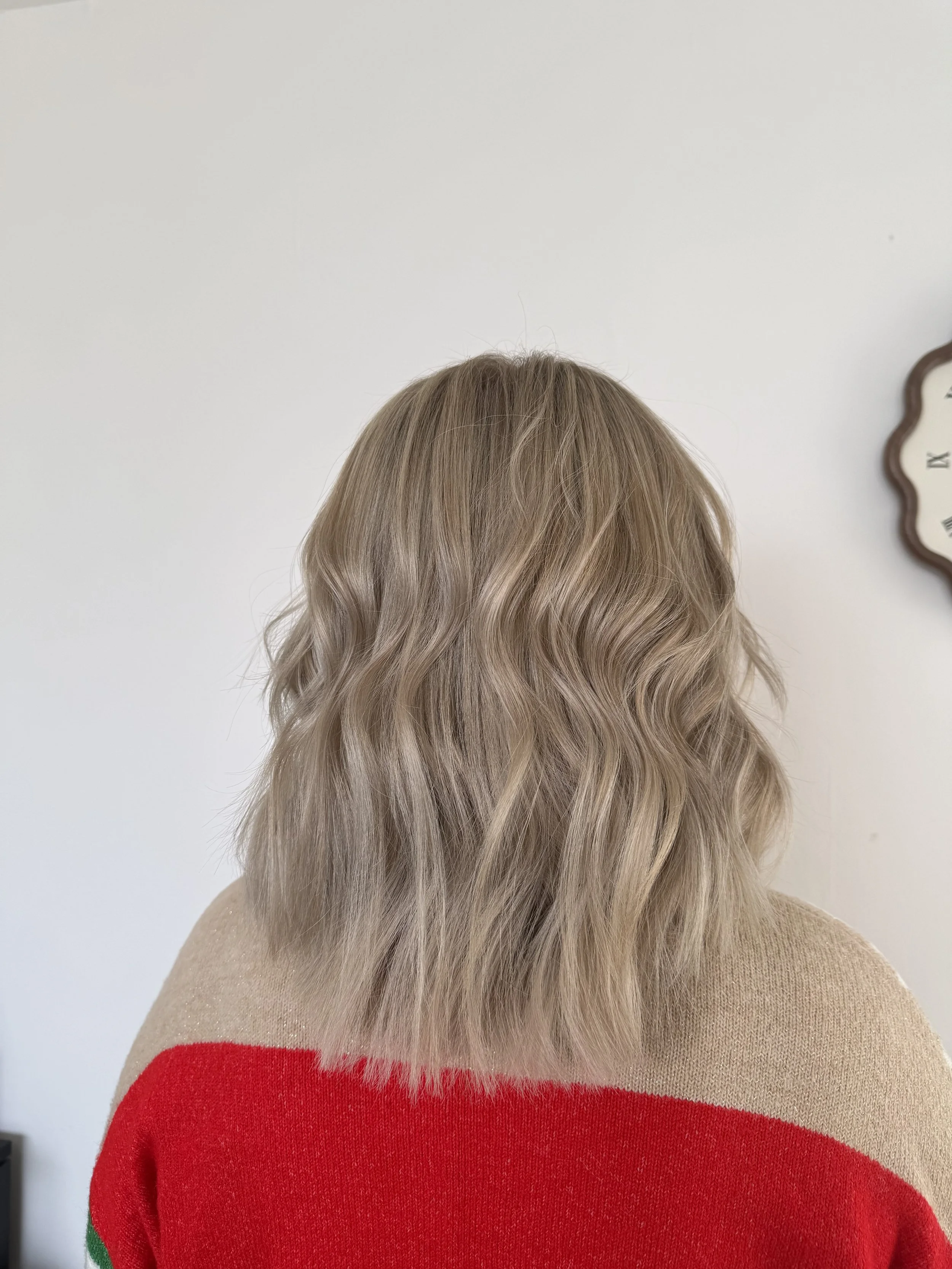 Back view of a woman with shoulder-length wavy blonde hair, wearing a multicolored sweater, against a white wall with a wall clock.