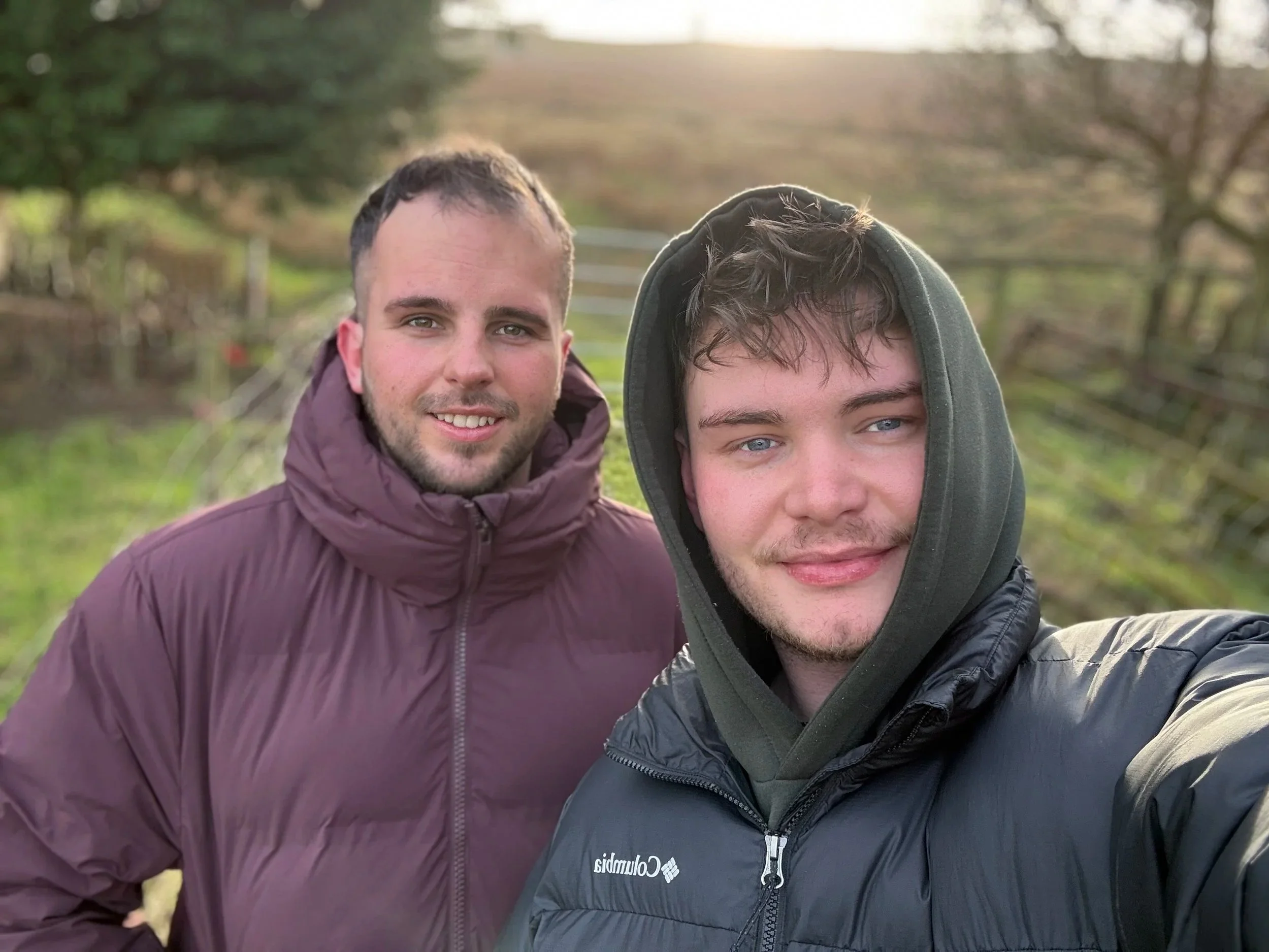 Two young men taking a selfie outdoors on a cloudy day, with trees and a fence in the background.