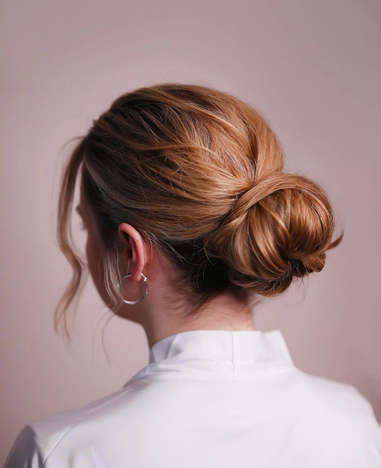 Back view of a woman with red hair styled in a low, twisted bun, wearing a white top and hoop earrings.