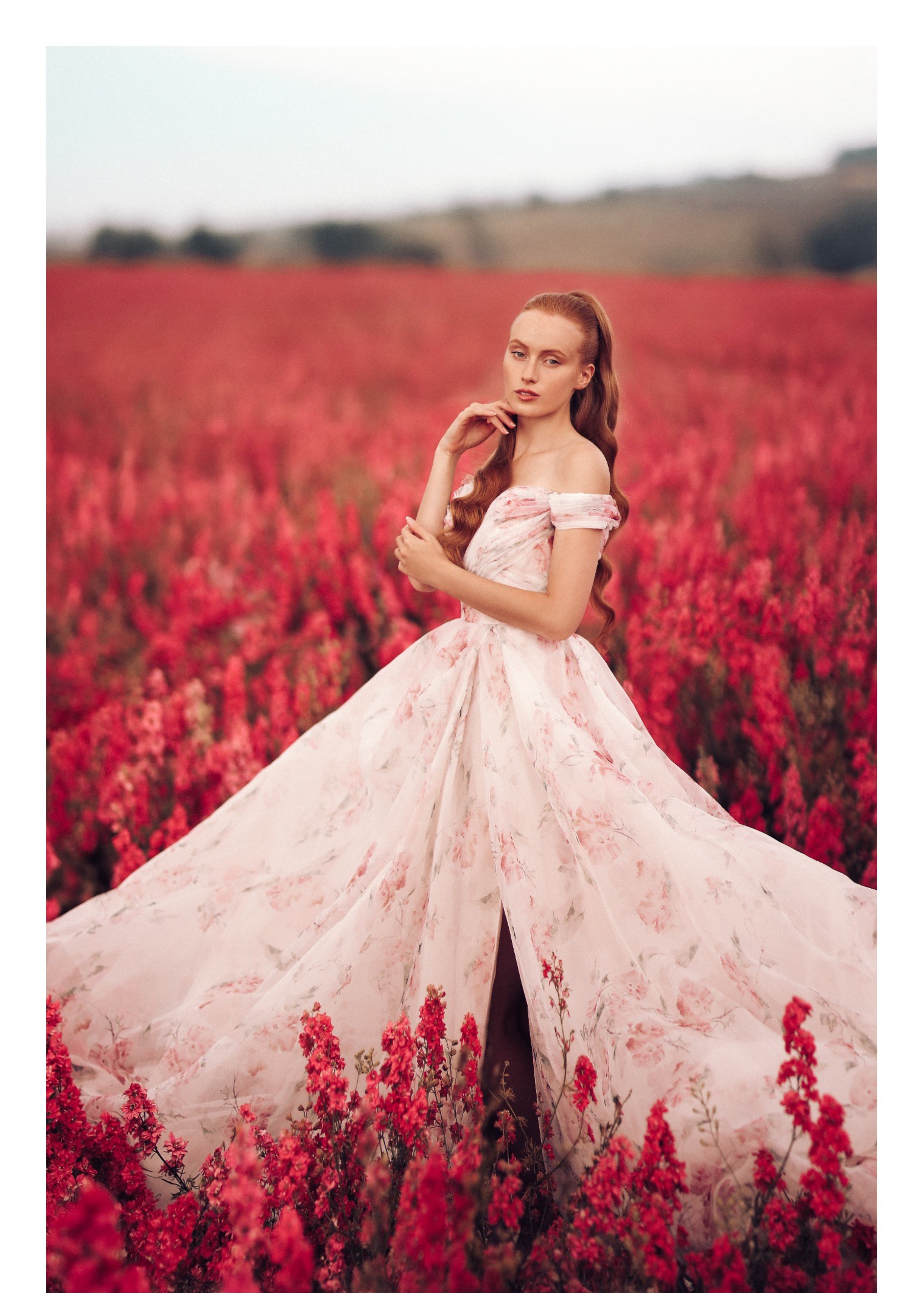 A woman in a flowing, off-shoulder pink floral dress standing in a field of pink flowers with a distant hillside in the background.