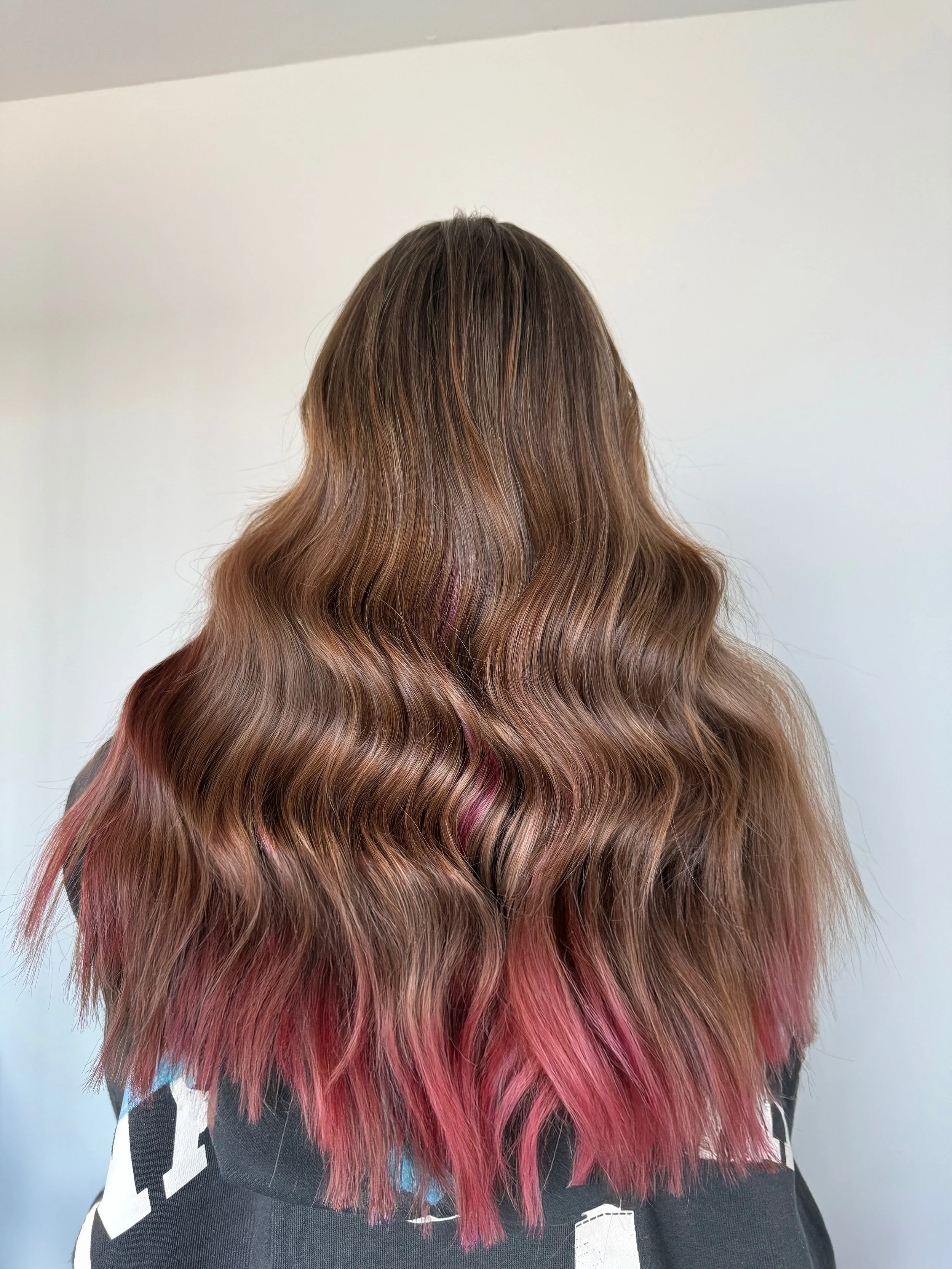 Back view of a woman with long, wavy hair that transitions from brown at the roots to pink at the tips.