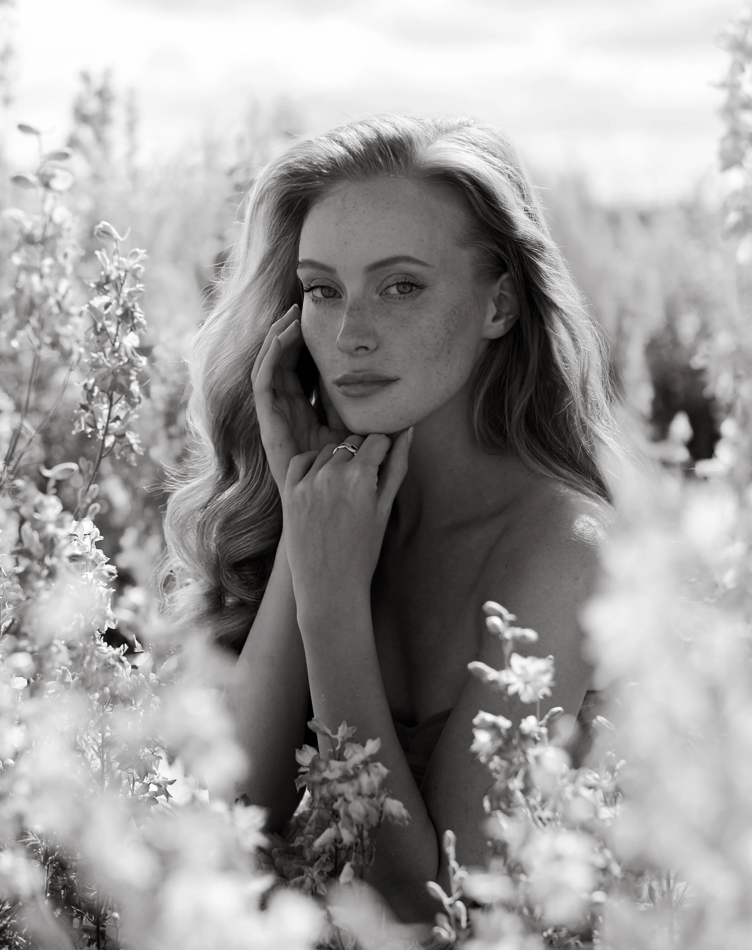 Black and white portrait of a young woman with wavy hair, surrounded by flowers, gazing into the camera with her hand near her face.