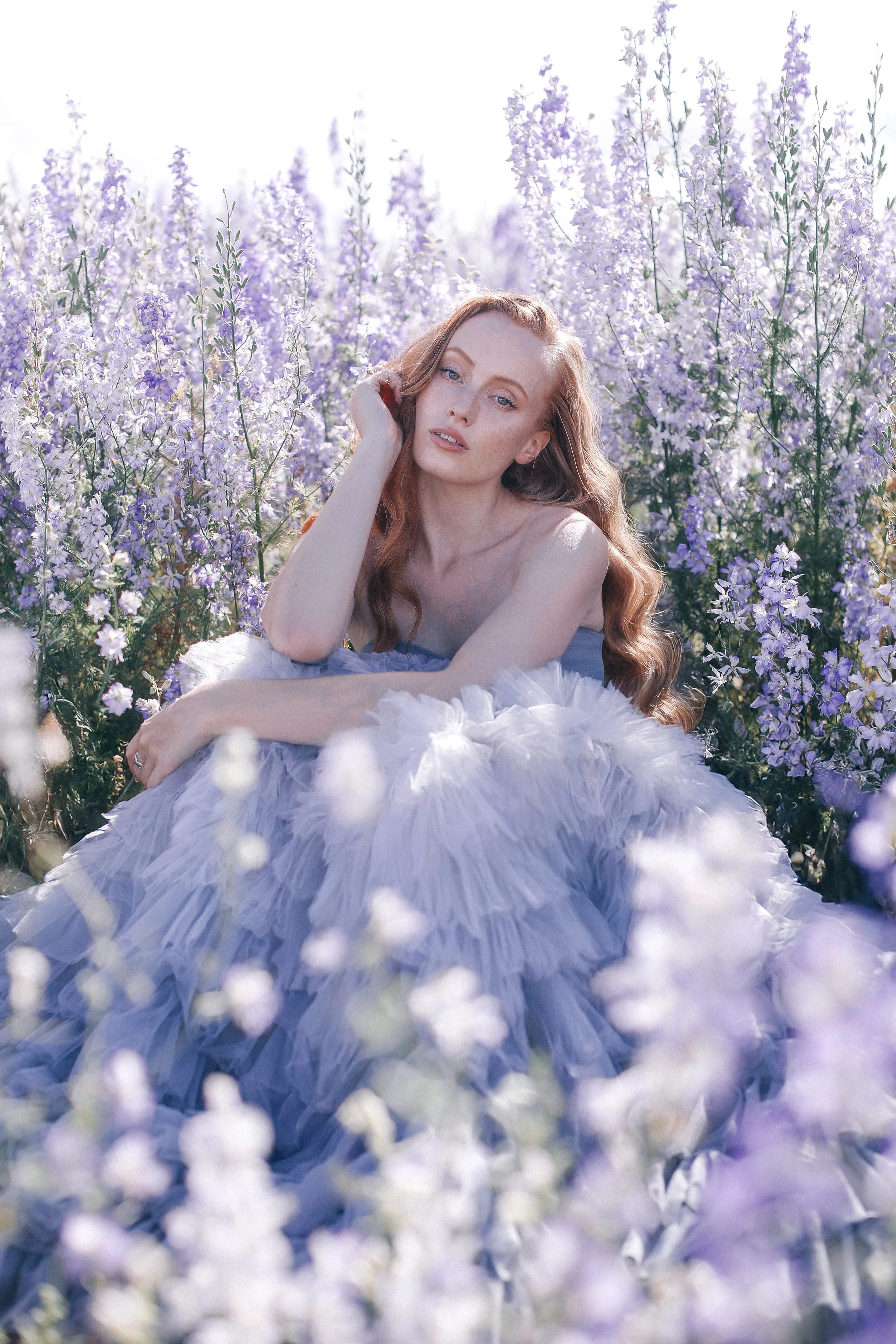 A woman with long red hair, wearing a pastel purple tulle gown, lying among lavender flowers in a sunlit field.