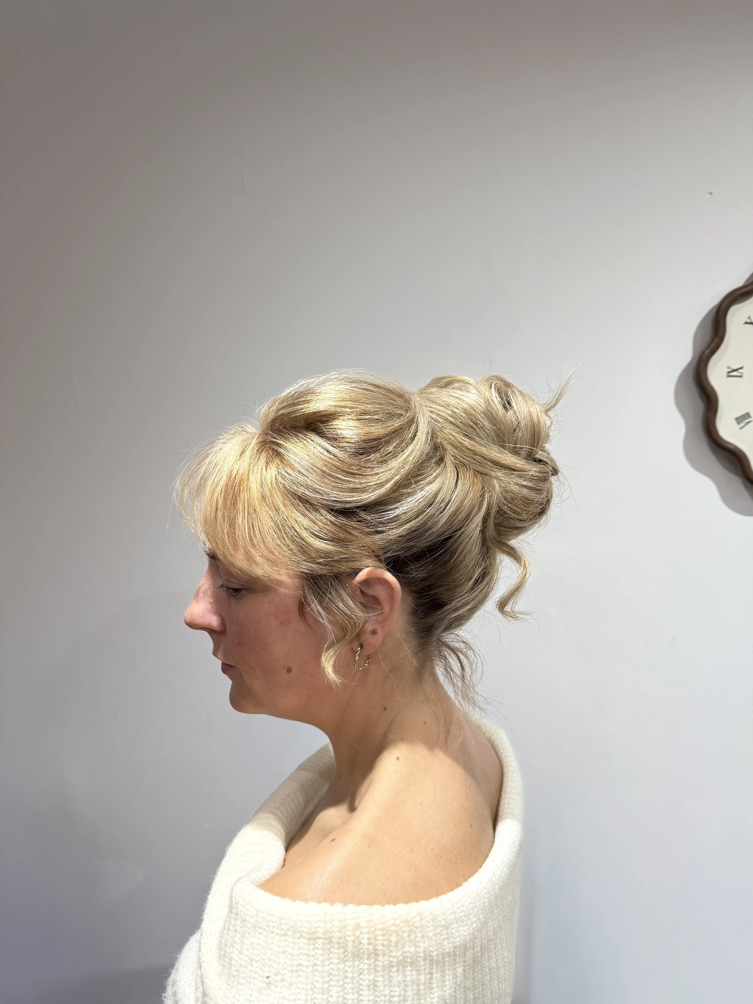 Side view of a woman with blonde, styled updo hair, wearing a white off-the-shoulder sweater, against a plain wall with a clock.