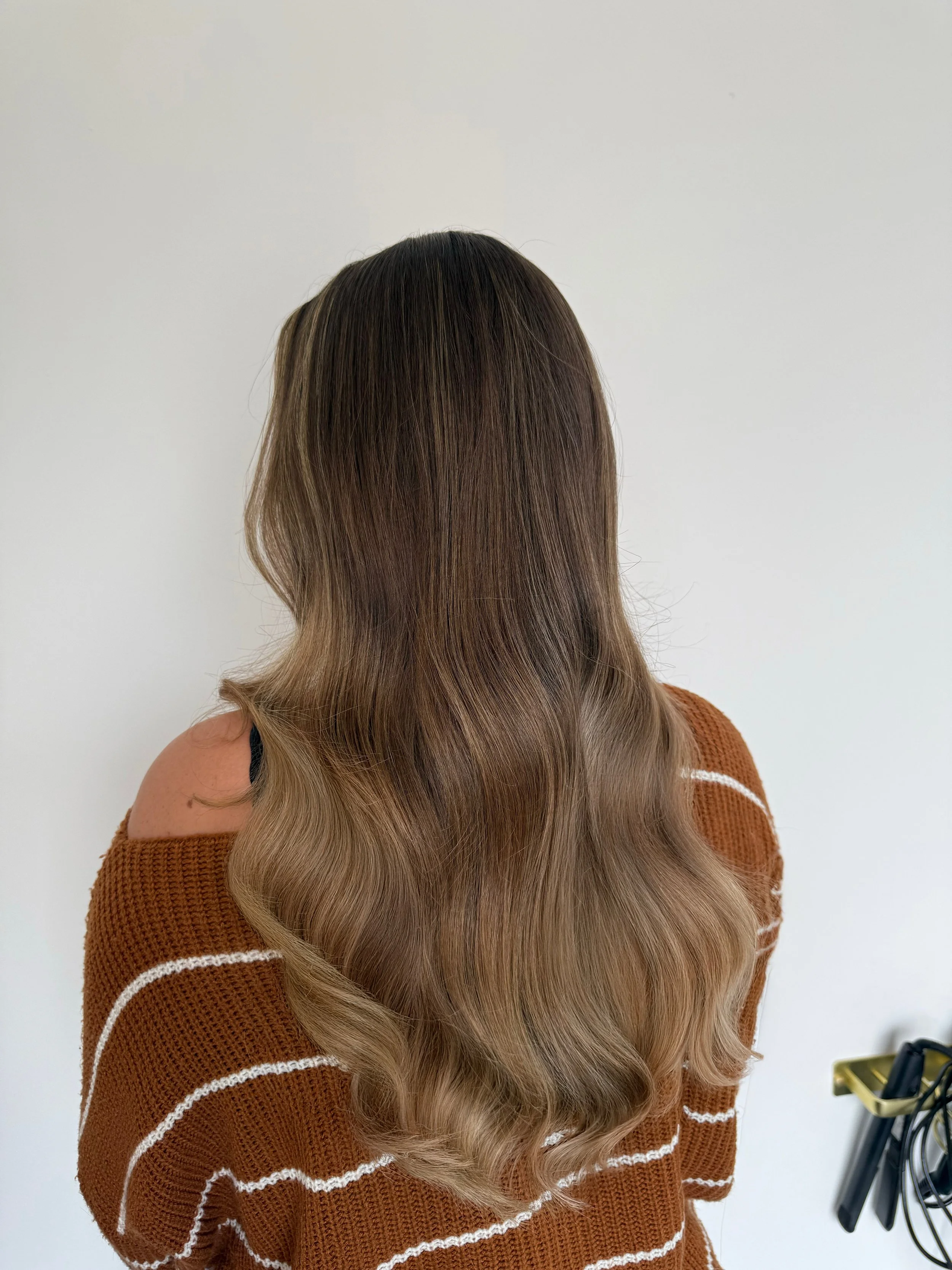 Back view of a woman with medium-length, wavy, brown hair wearing an orange striped sweater, standing against a plain white wall with some cords on the right side.