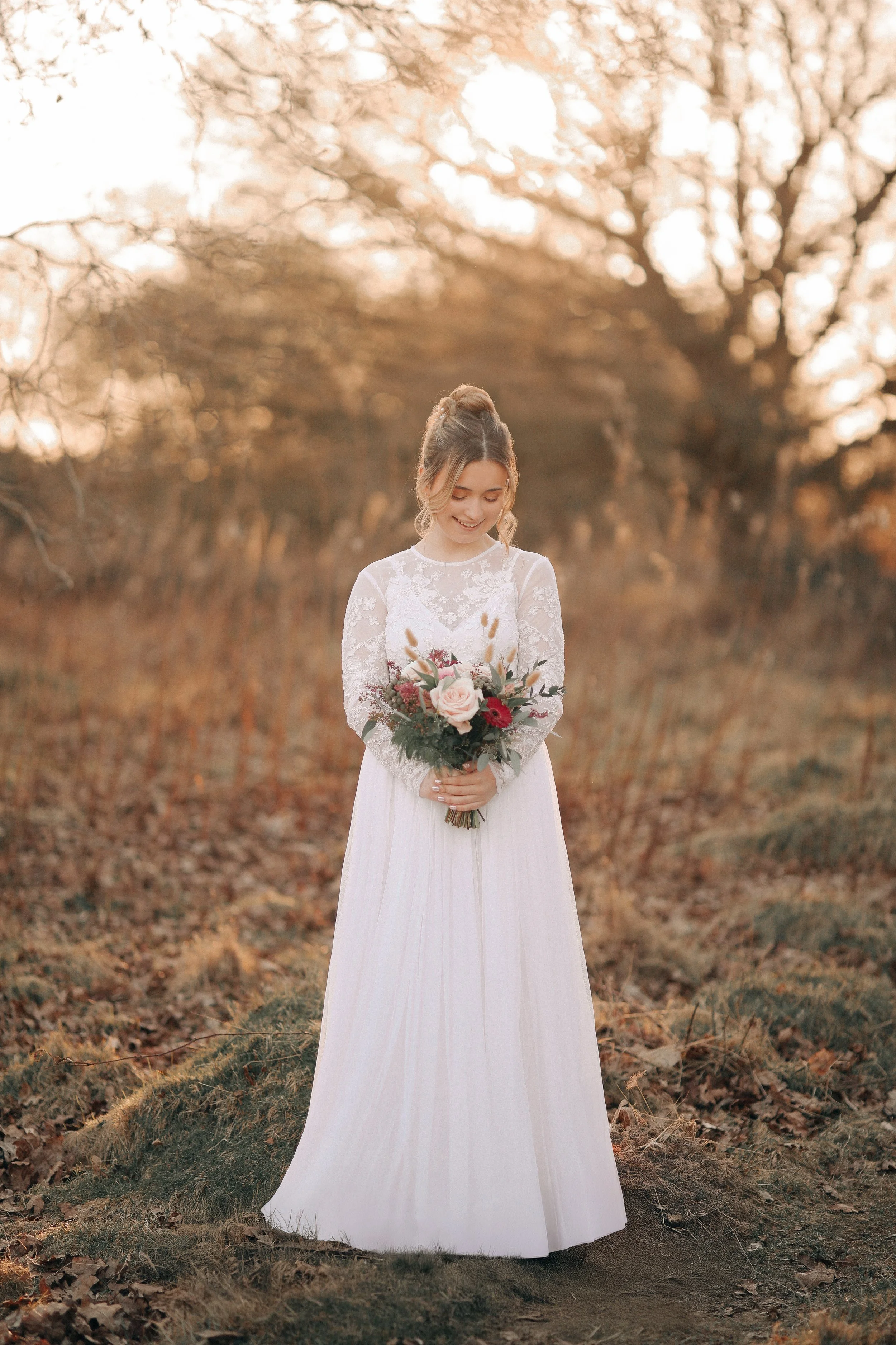 A bride in a white lace wedding dress holding a colorful bouquet, standing outdoors on a natural, wooded background during autumn.