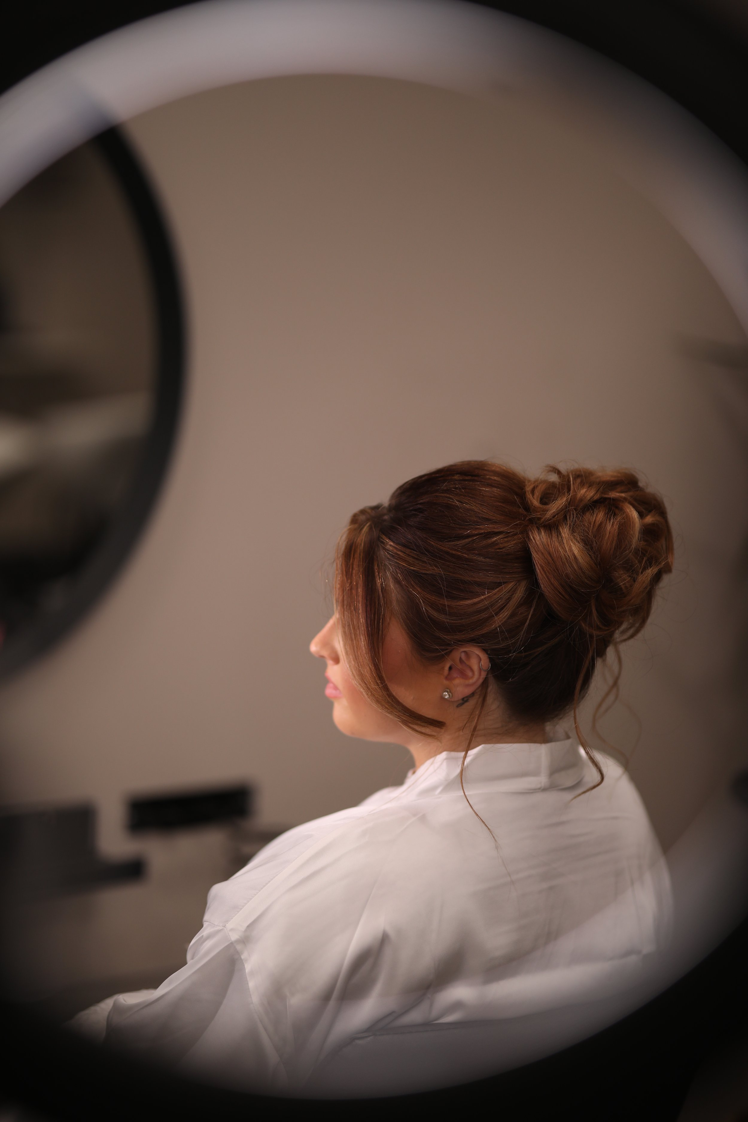 A woman with styled brown hair in an updo, wearing pearl earrings and a white robe, seen through a circular mirror or lens.