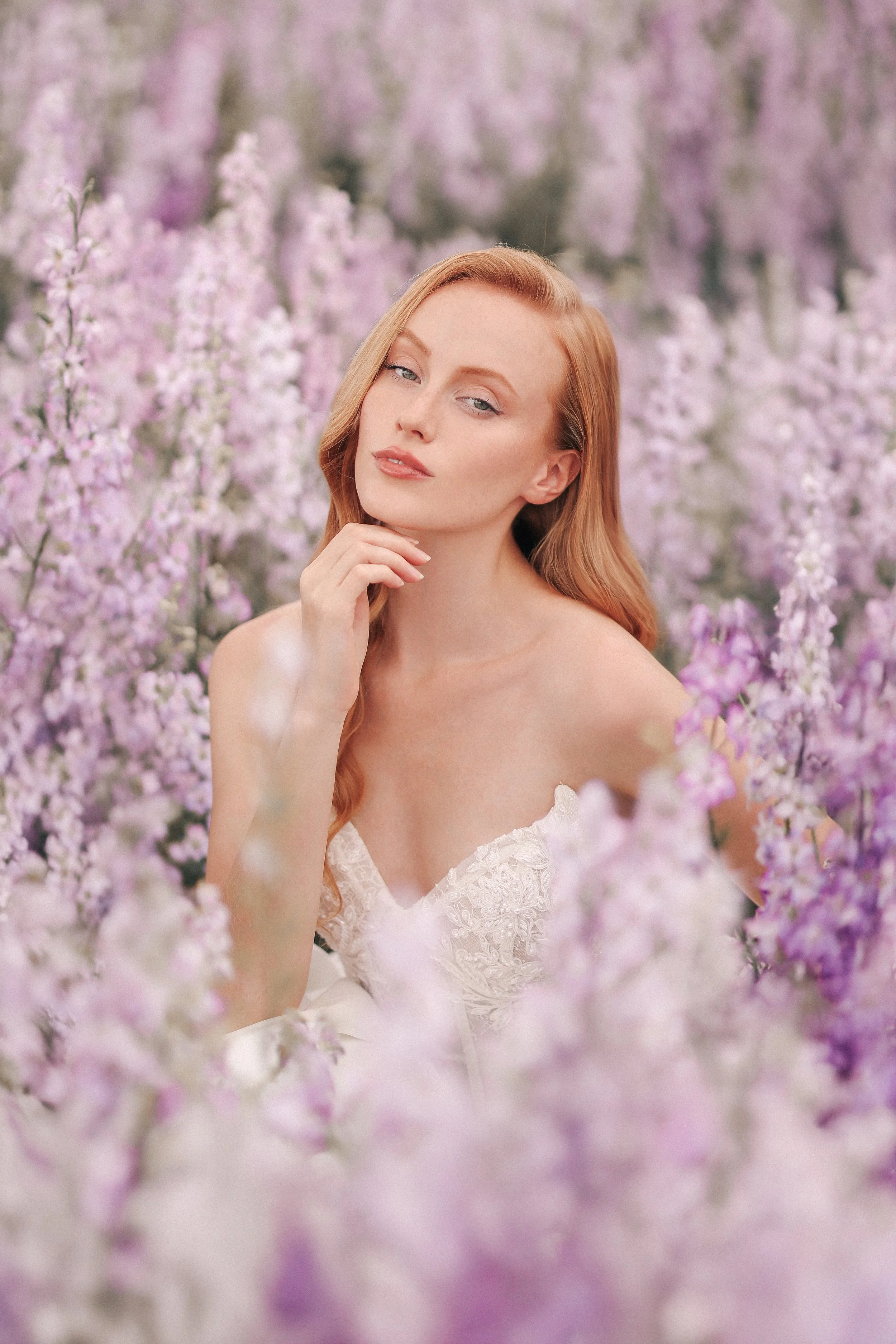 A woman with red hair wearing a white lace dress, surrounded by pink and purple flowers in a field.