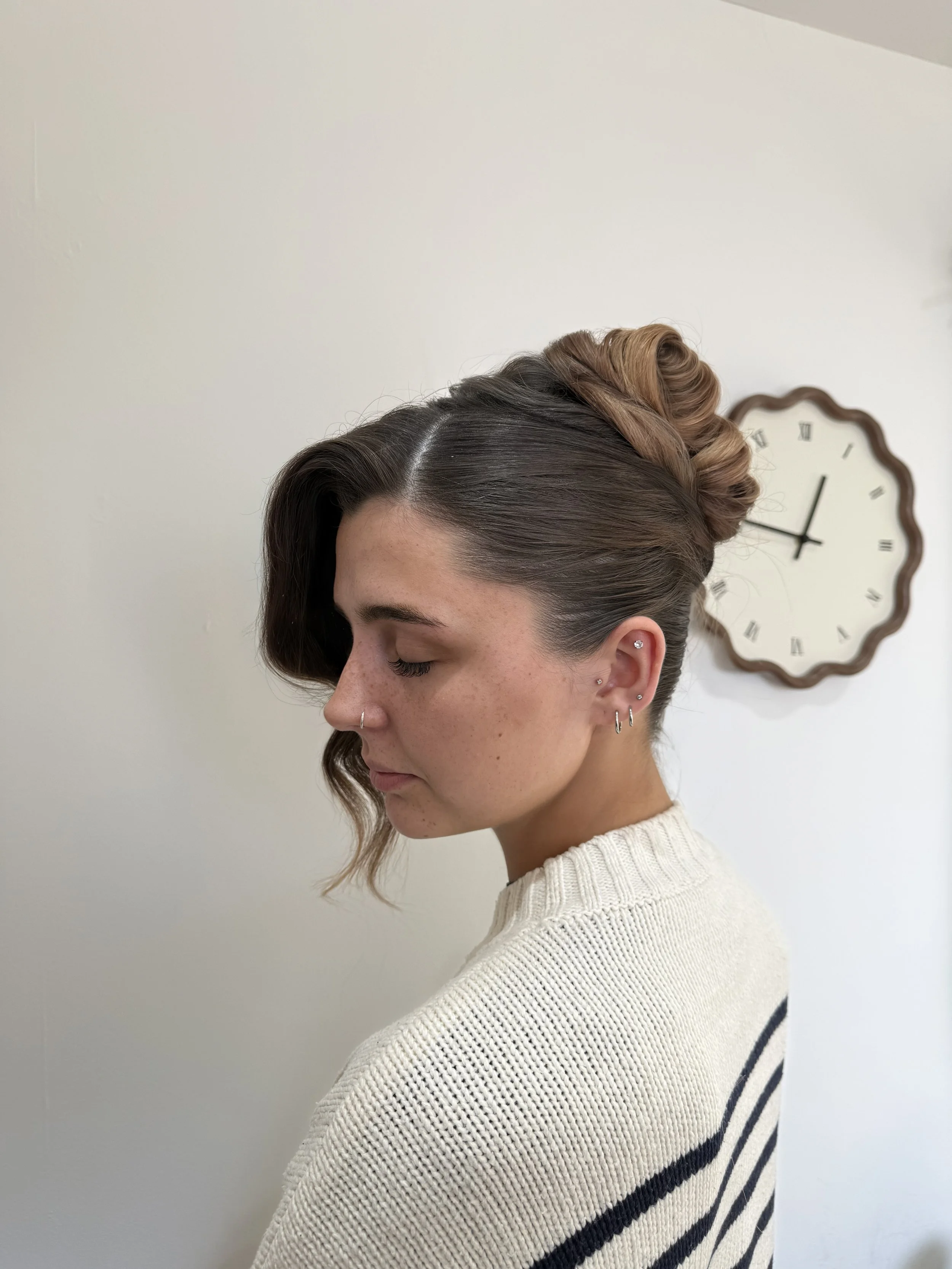 Side profile of a woman with vintage-style curled hair pinned up, wearing a cream sweater with black stripes, standing in front of a white wall with a clock.