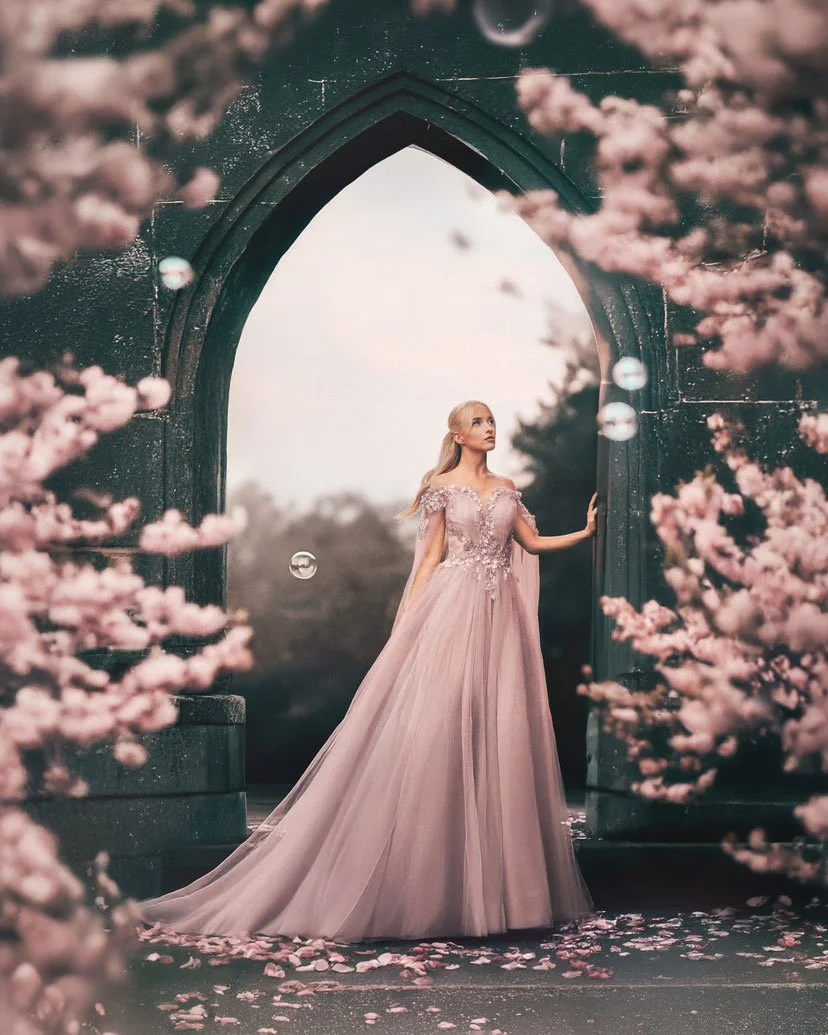 A woman in a flowing pink gown standing under an arched stone structure, surrounded by pink cherry blossom trees.