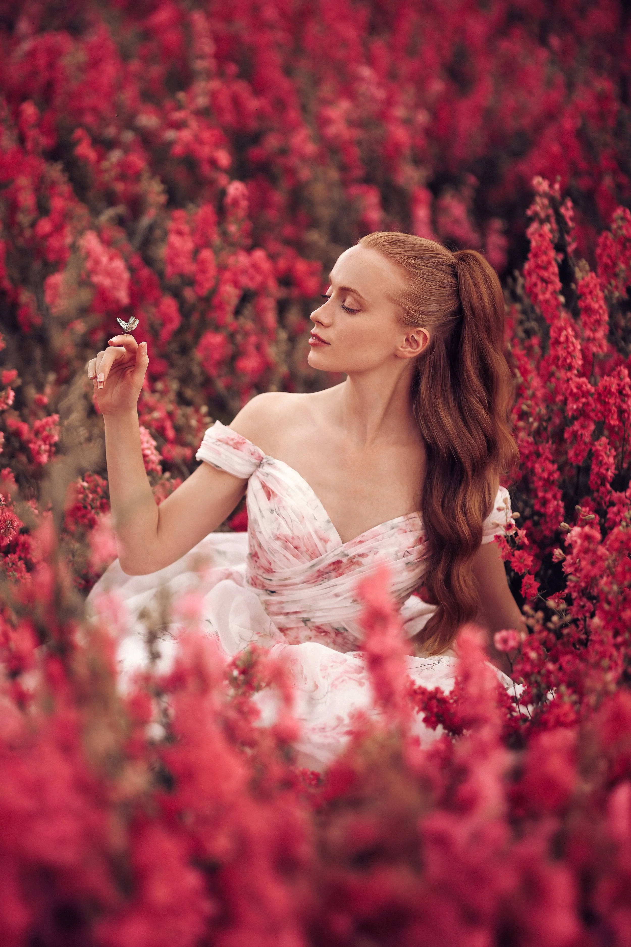 A woman in a pink floral dress sitting in a field of pink flowers, holding a butterfly on her finger.