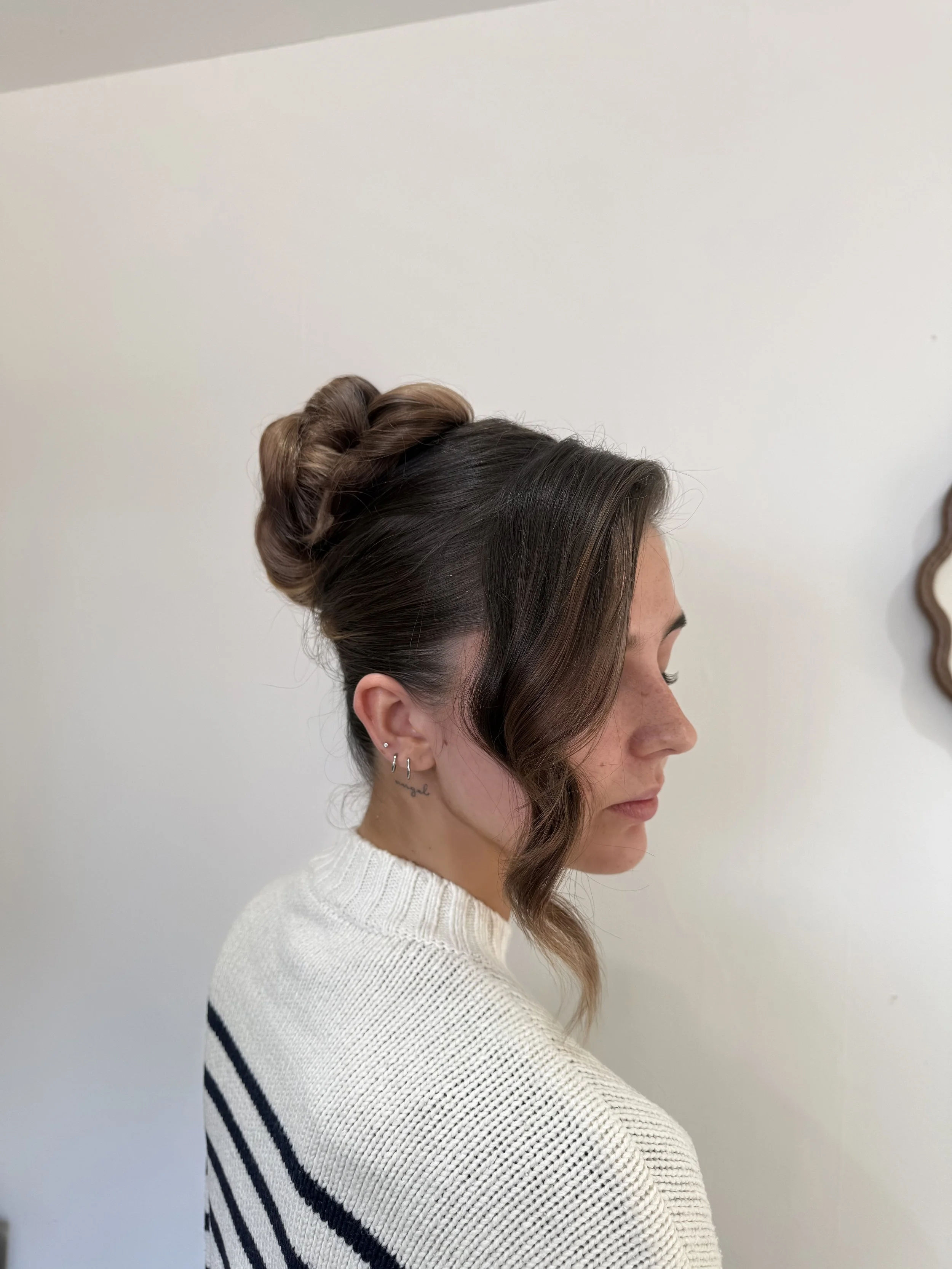 Side profile of a woman with styled hair in an updo and a loose tendril, wearing a white knit sweater with black stripes.