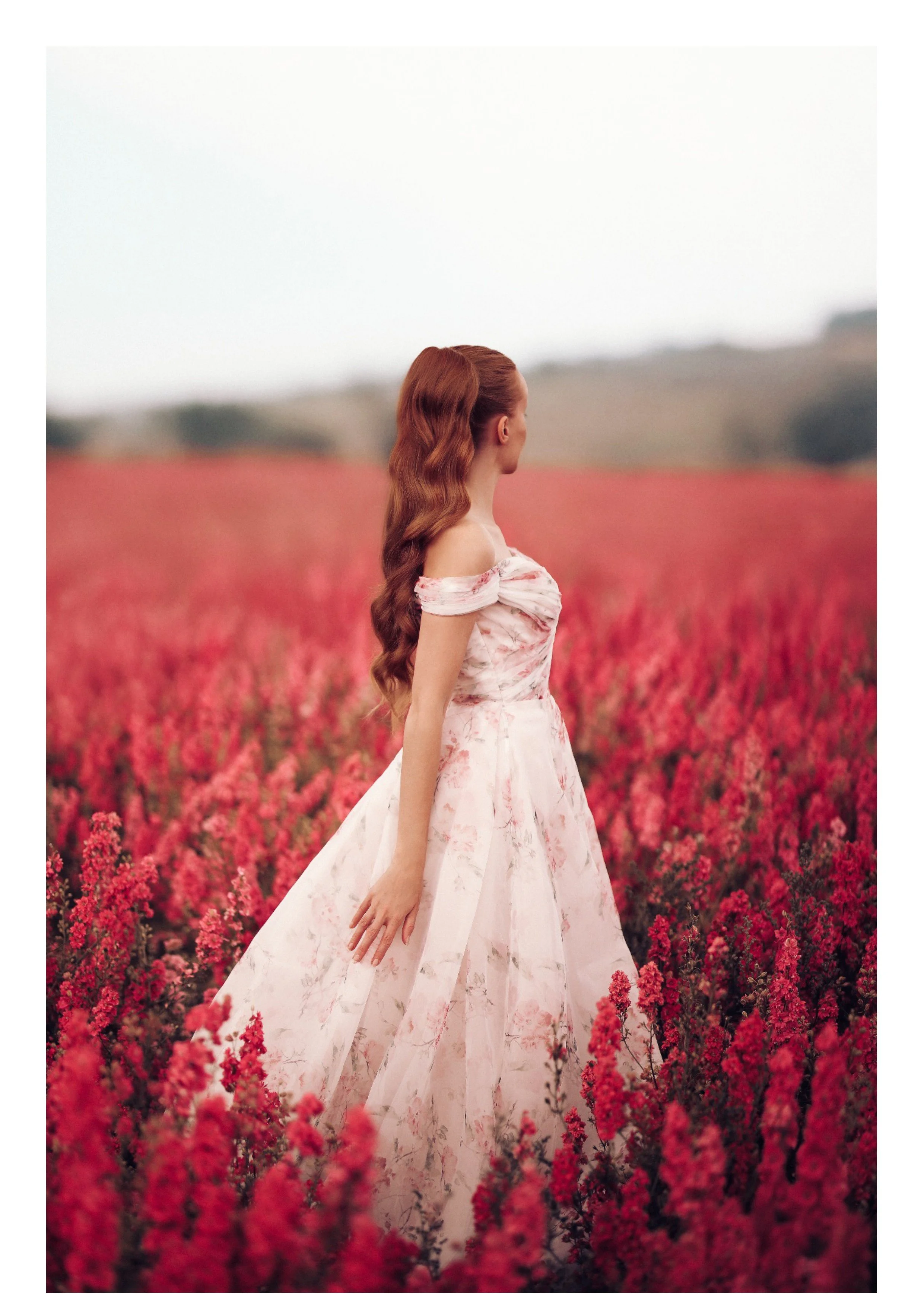 A woman with long, wavy red hair in a pink floral off-shoulder dress standing in a field of pink flowers, facing away and looking to the right.