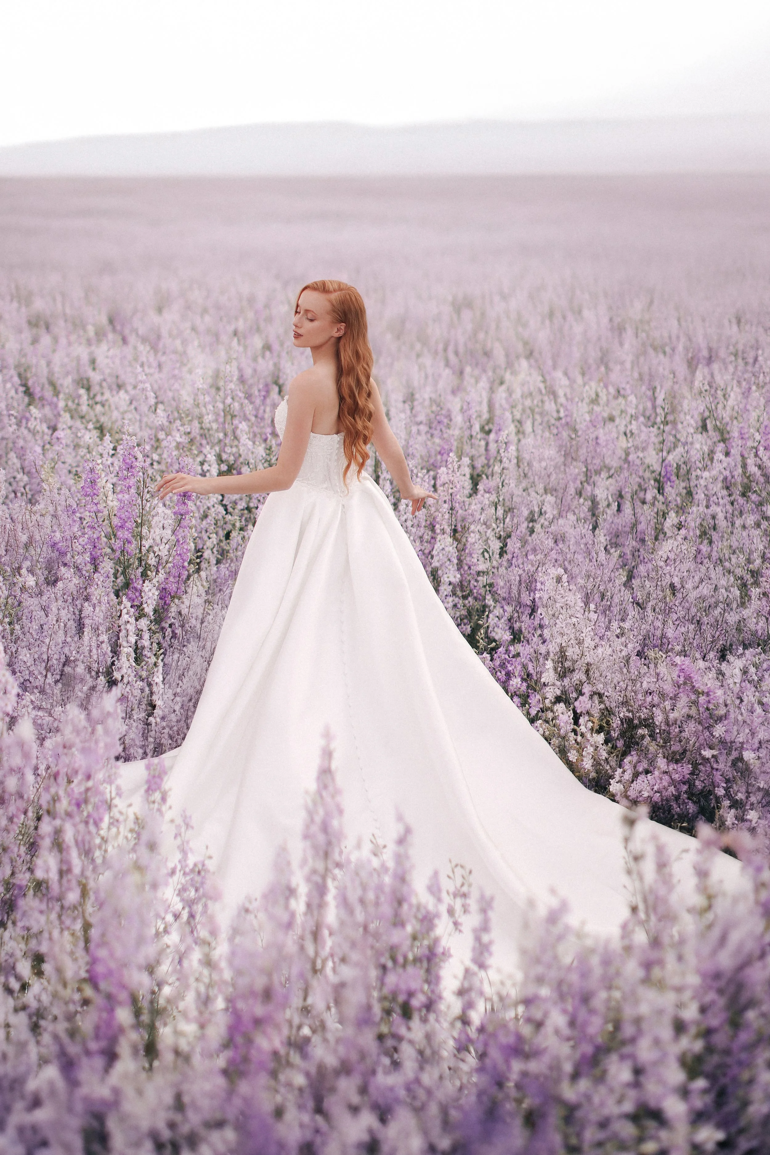 A woman in a white wedding dress standing in a lavender field, with her eyes closed and long red hair flowing.