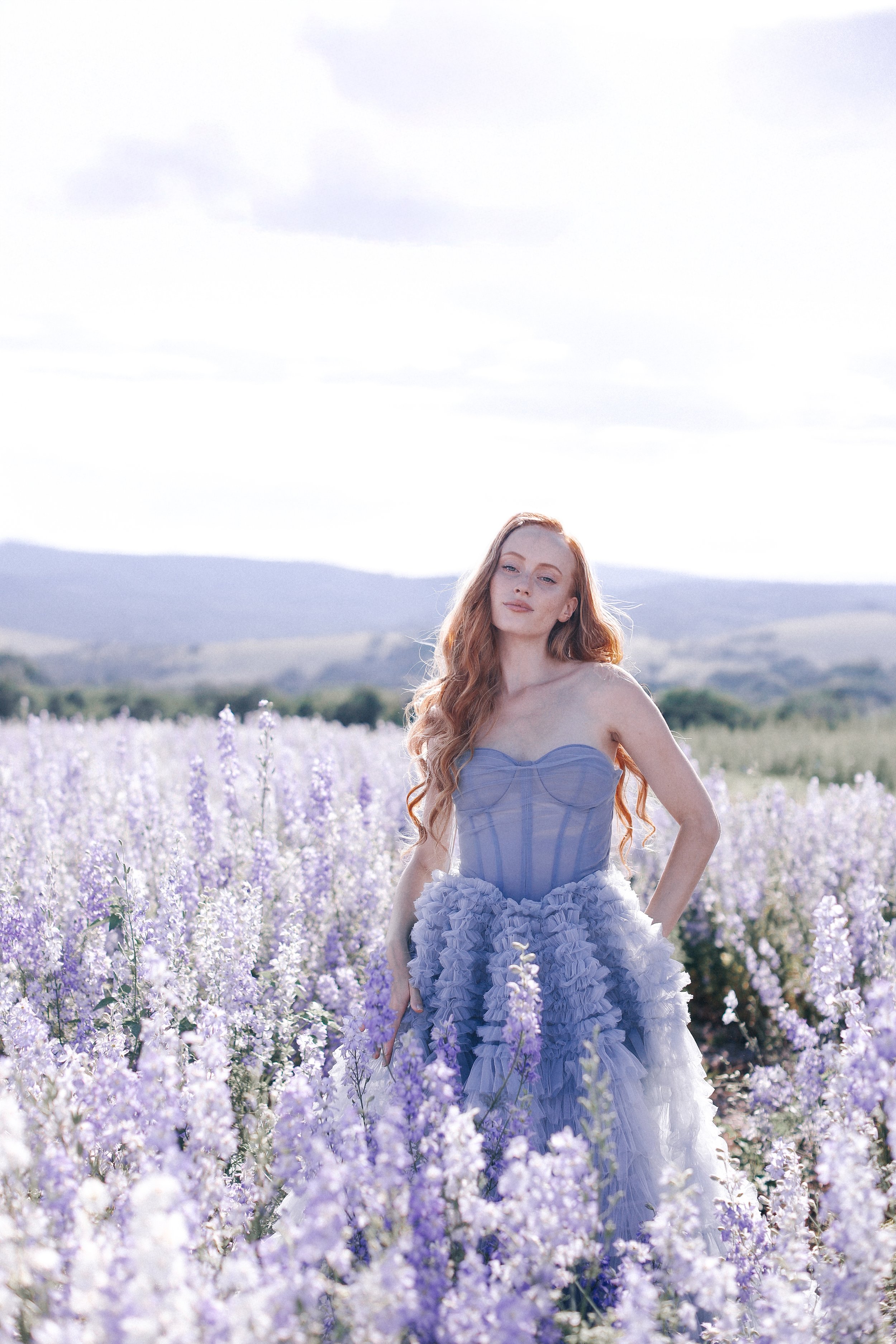 A woman in a lavender dress standing in a field of blooming lavender flowers, with hills in the background under a cloudy sky.