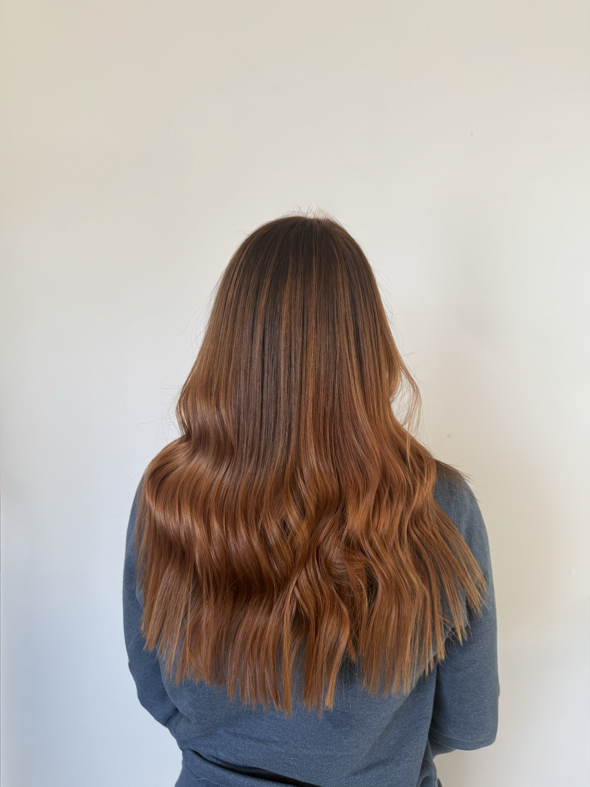 Back view of a woman with long, wavy, light brown hair standing against a plain white wall.