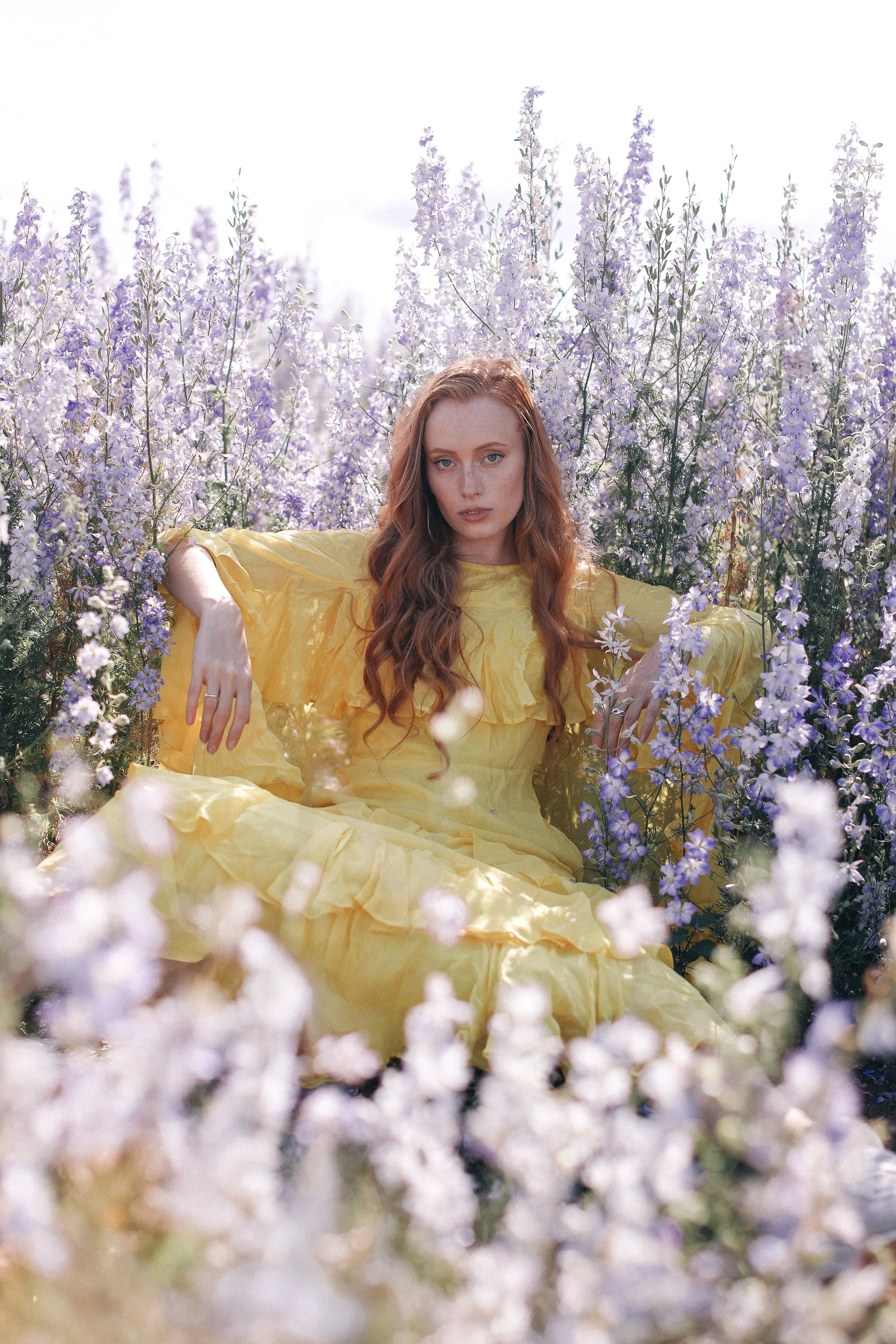 A woman with long red hair wearing a yellow dress sitting among purple flowers.