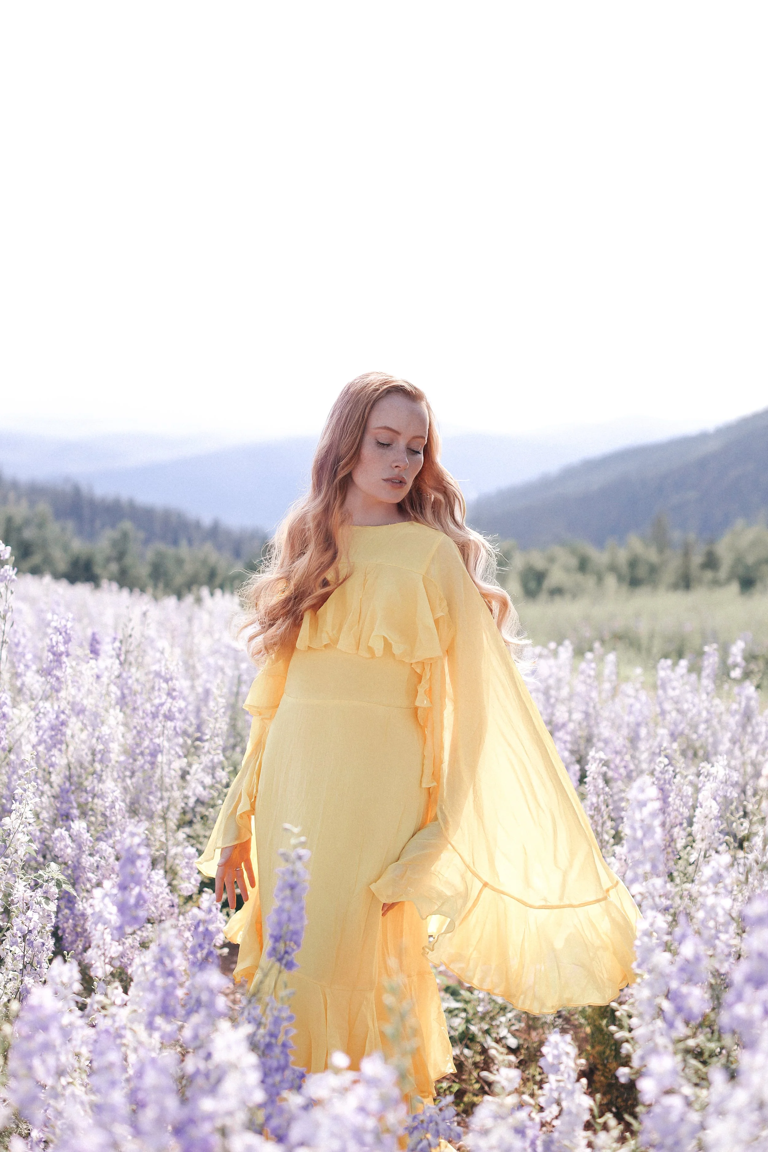 A woman with long, wavy, red hair standing in a field of purple flowers, wearing a flowing yellow dress with ruffled details, with mountains and a cloudy sky in the background.