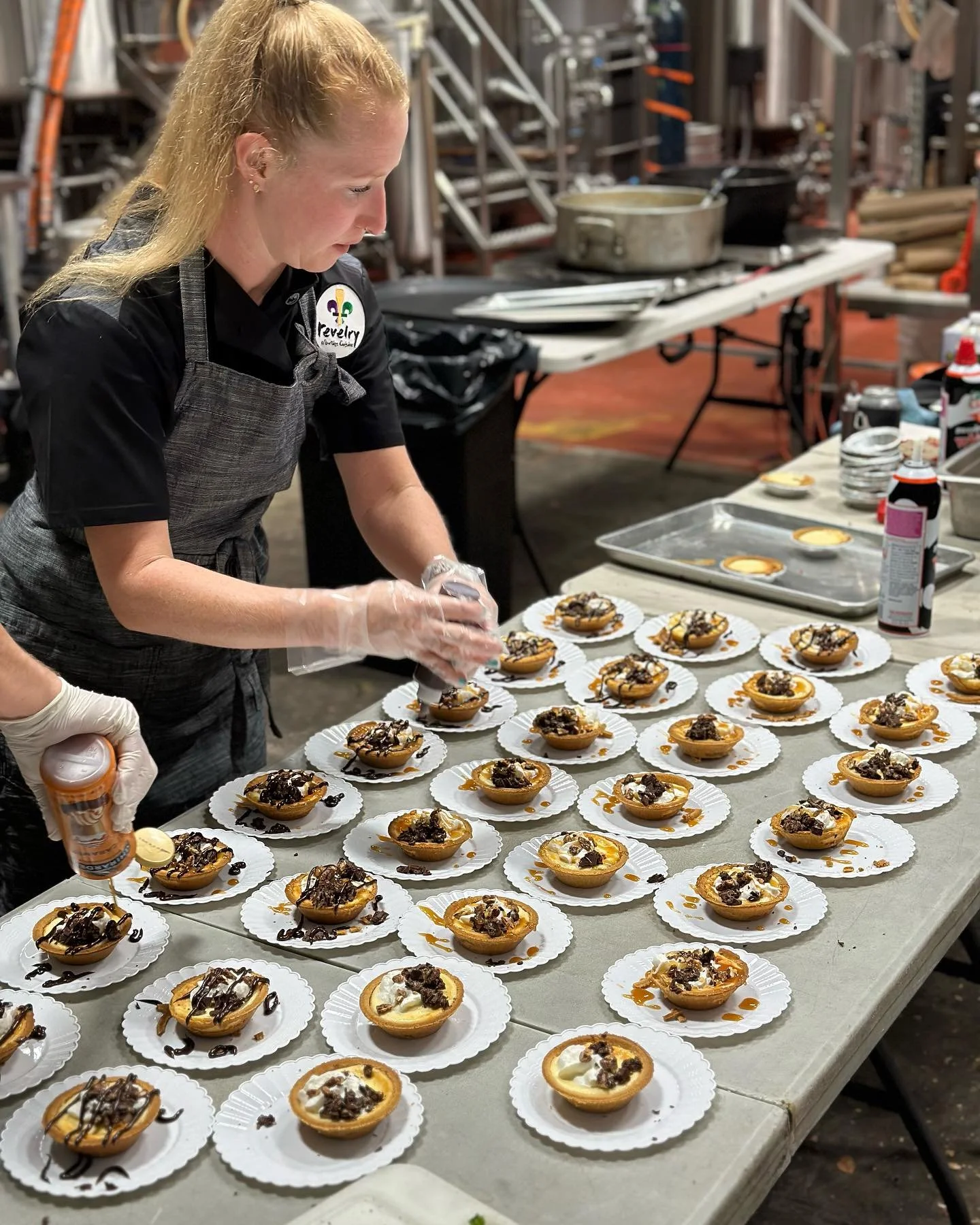 A woman preparing small desserts topped with cream and chocolate on a table in a kitchen or bakery setting.