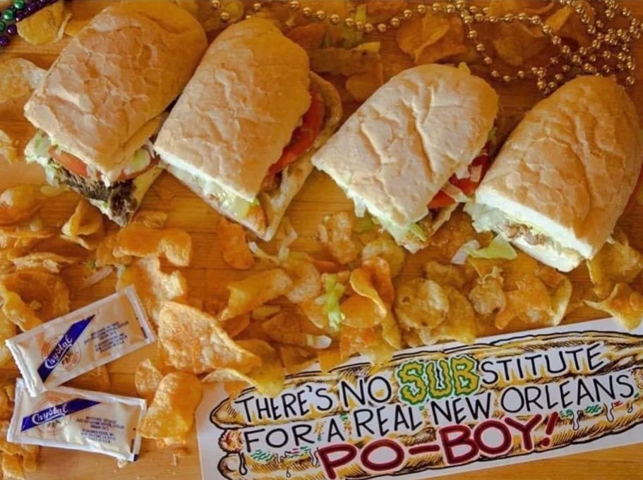 Four po' boy sandwiches on a wooden surface surrounded by potato chips, Mardi Gras beads, and packets of Crystal hot sauce. Sign reads: "There's no substitute for a real New Orleans po' boy!"