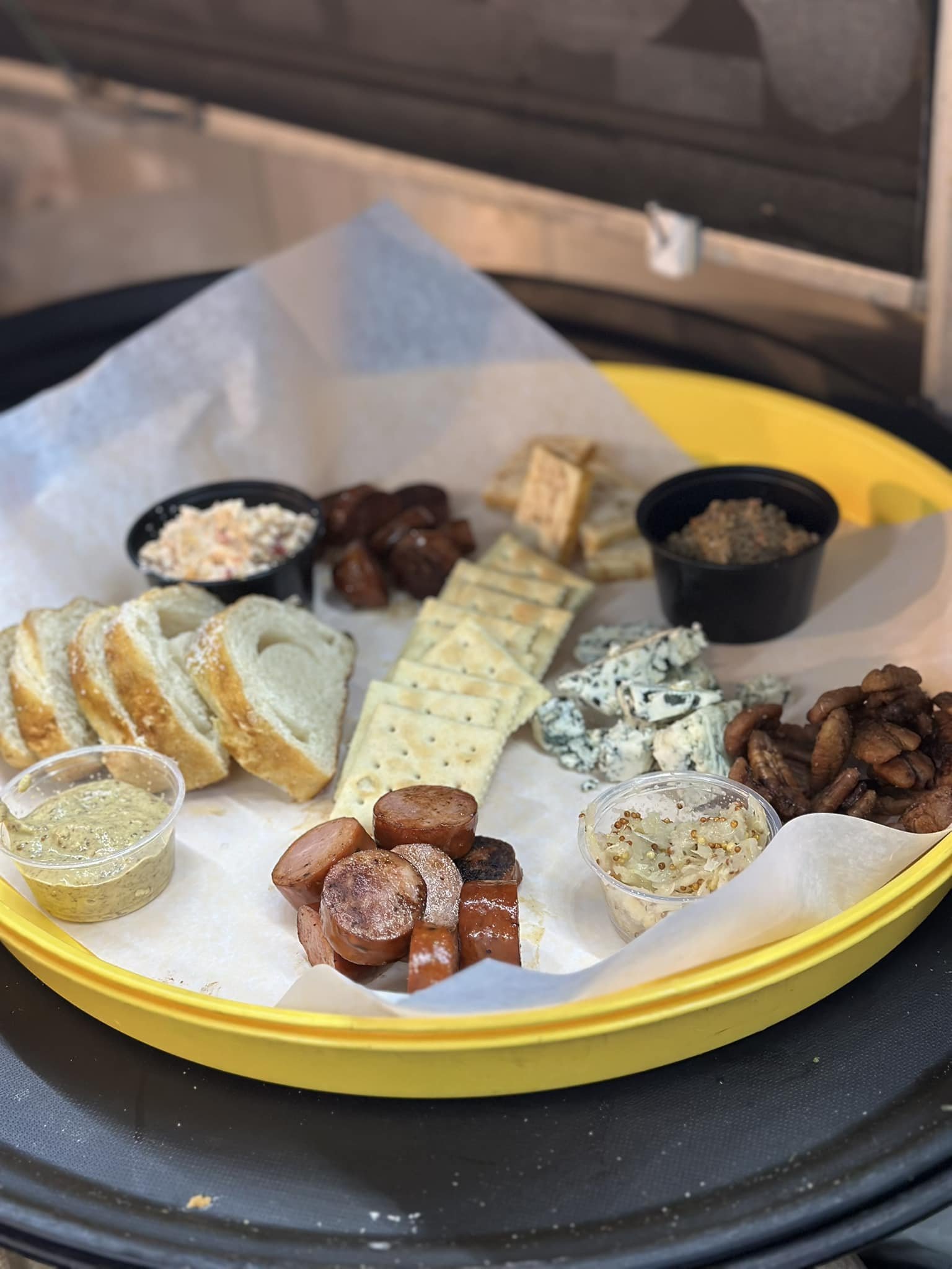 Charcuterie board with sliced bread, crackers, sausages, blue cheese, dips, and nuts on a yellow platter.