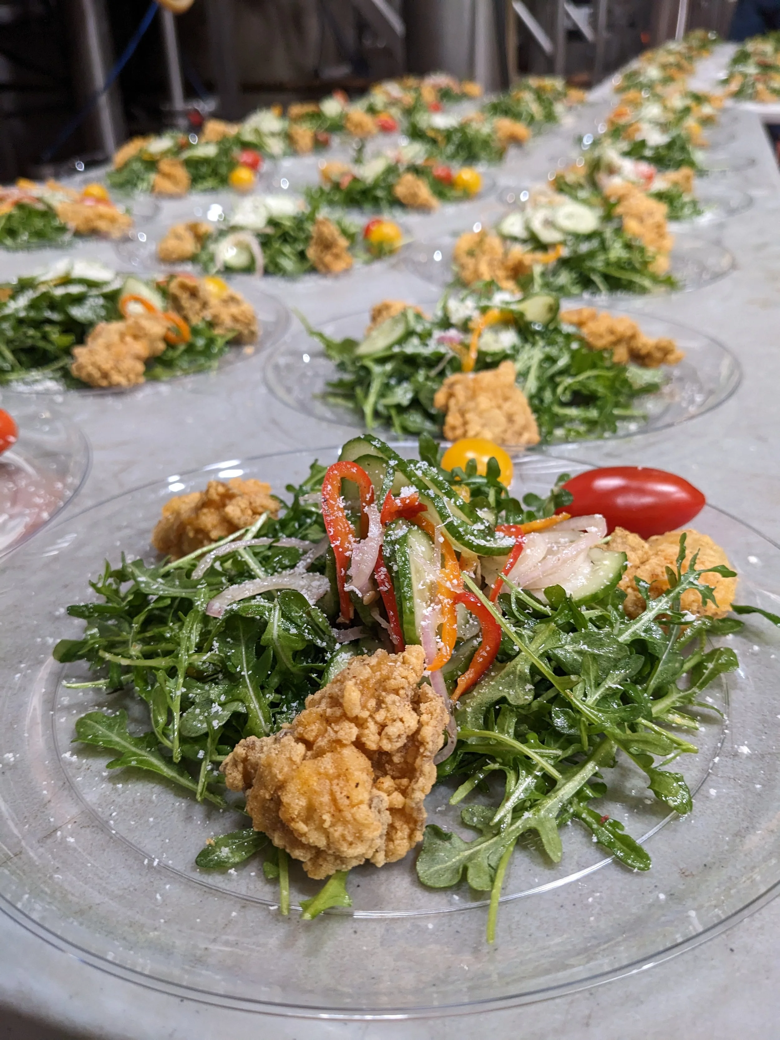 Multiple plates of fried chicken salads with arugula, sliced cucumbers, cherry tomatoes, shredded carrots, red onions, and sprinkled with grated cheese, lined up on a table.