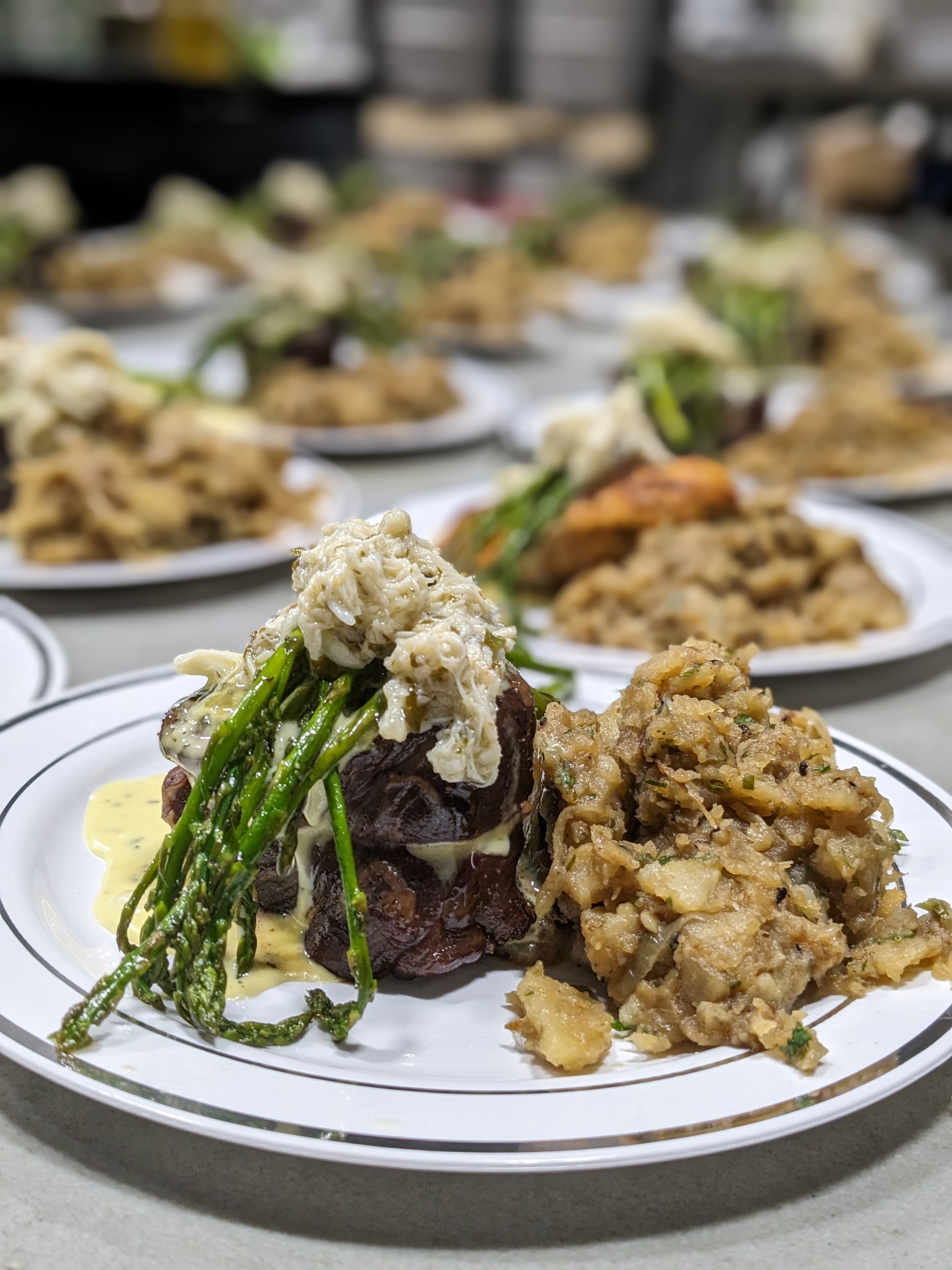 A plated dish with pieces of meat topped with asparagus, crab meat, served with a side of potato salad, on a white plate with gold trim.