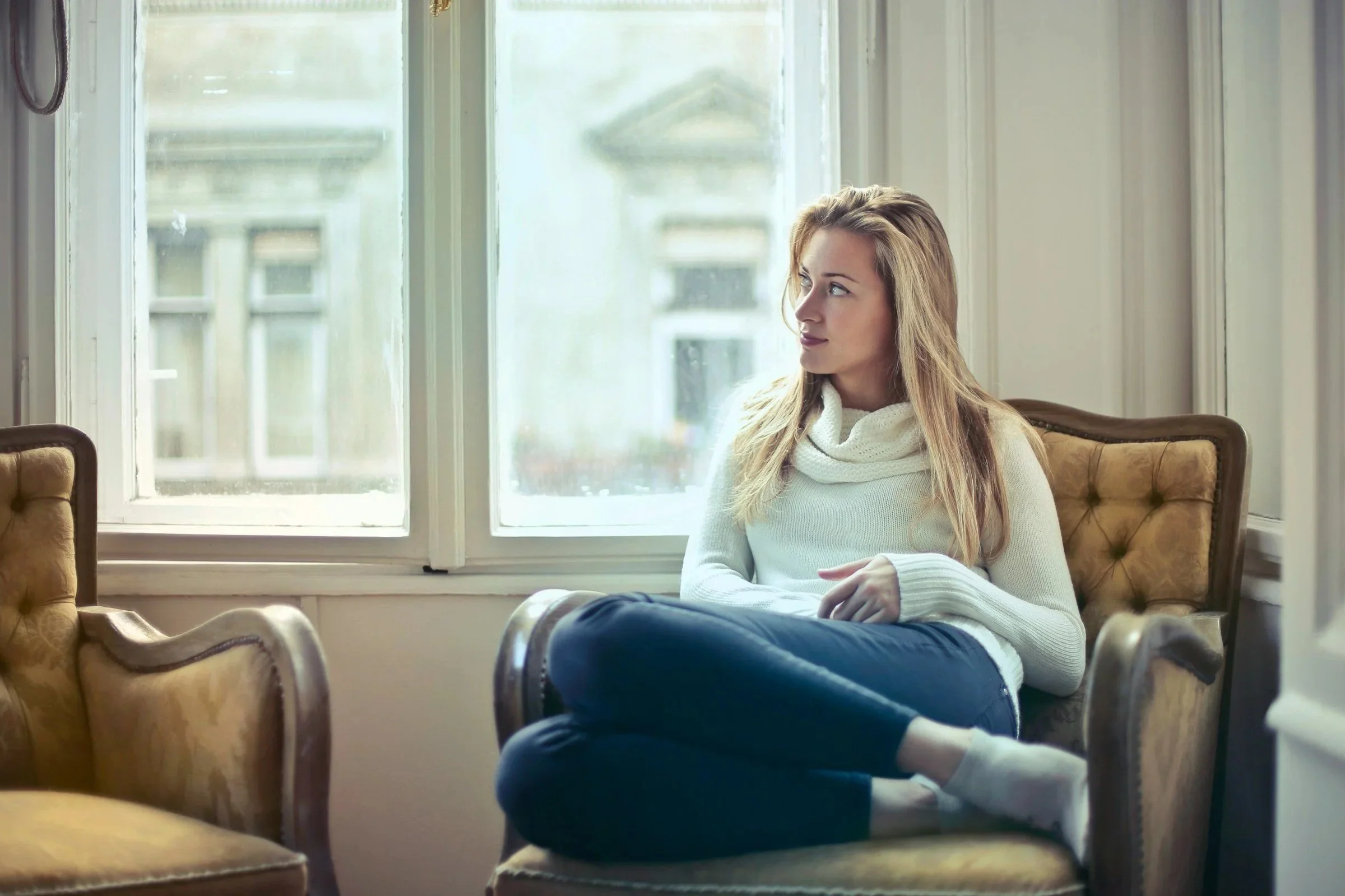A young woman with long blond hair sitting by a large window, wearing a cream sweater and navy jeans, looking thoughtfully outside.