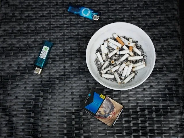 A white bowl filled with cigarette butts and ash on a black woven surface, surrounded by cigarette packs and lighters.