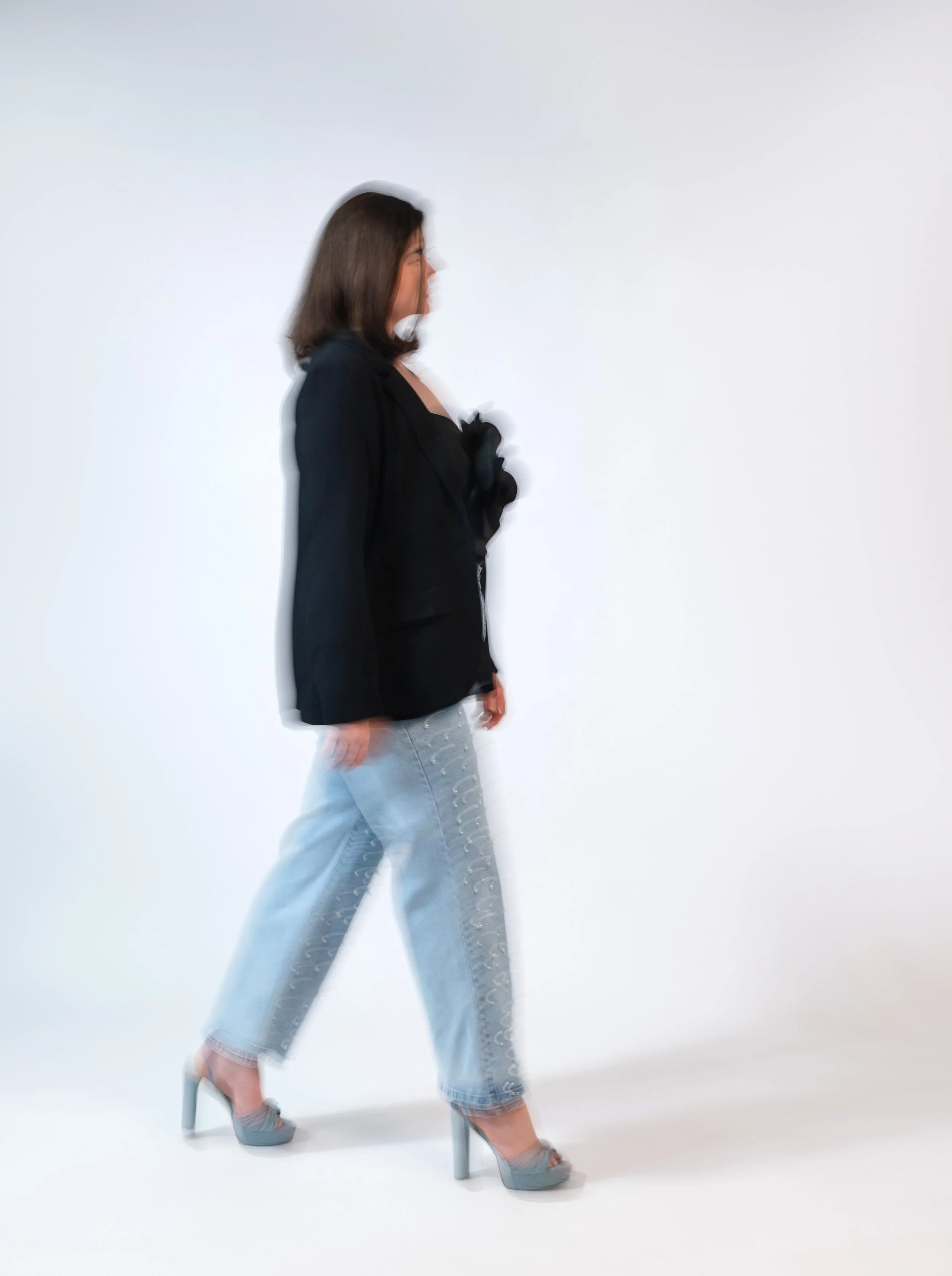 A woman walking in front of a plain white background, dressed in a black blazer, light-wash jeans with embellishments, and high heels.