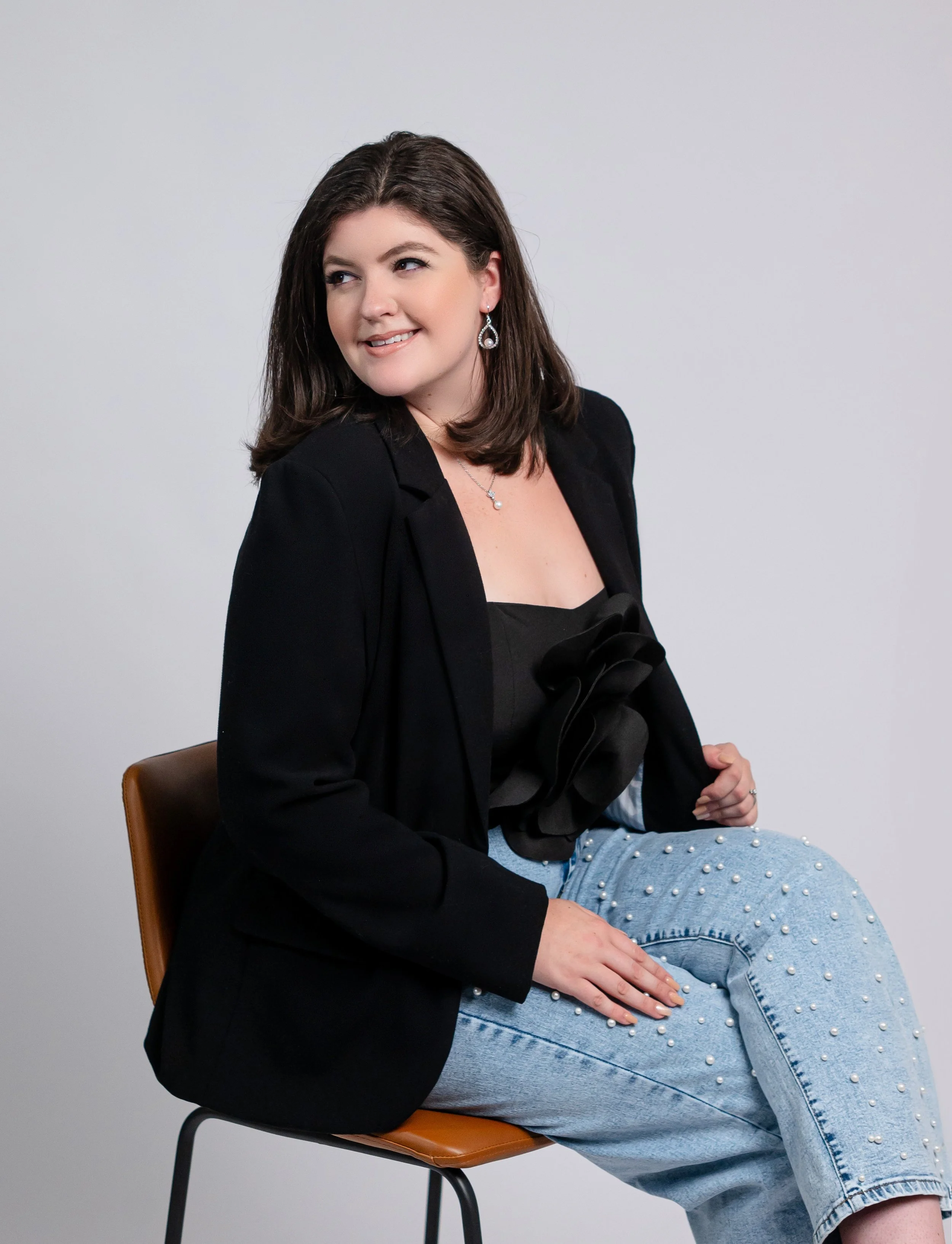A woman with shoulder-length dark brown hair smiling while seated on a brown chair against a plain light gray background. She is wearing a black blazer, black top with a large decorative bow, light blue jeans with pearl embellishments, earrings, and a necklace.