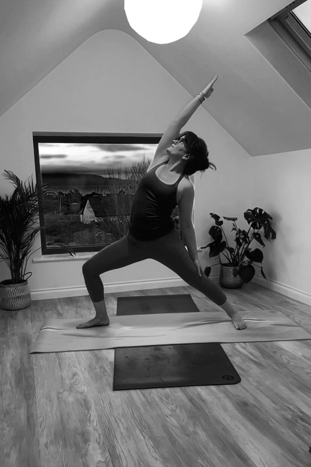 A woman practicing yoga in a room with a sloped ceiling, wooden floor, and potted plants. She is in a warrior pose, standing on a yoga mat with her right arm extended upward and her left hand on her left thigh, looking towards her raised hand.