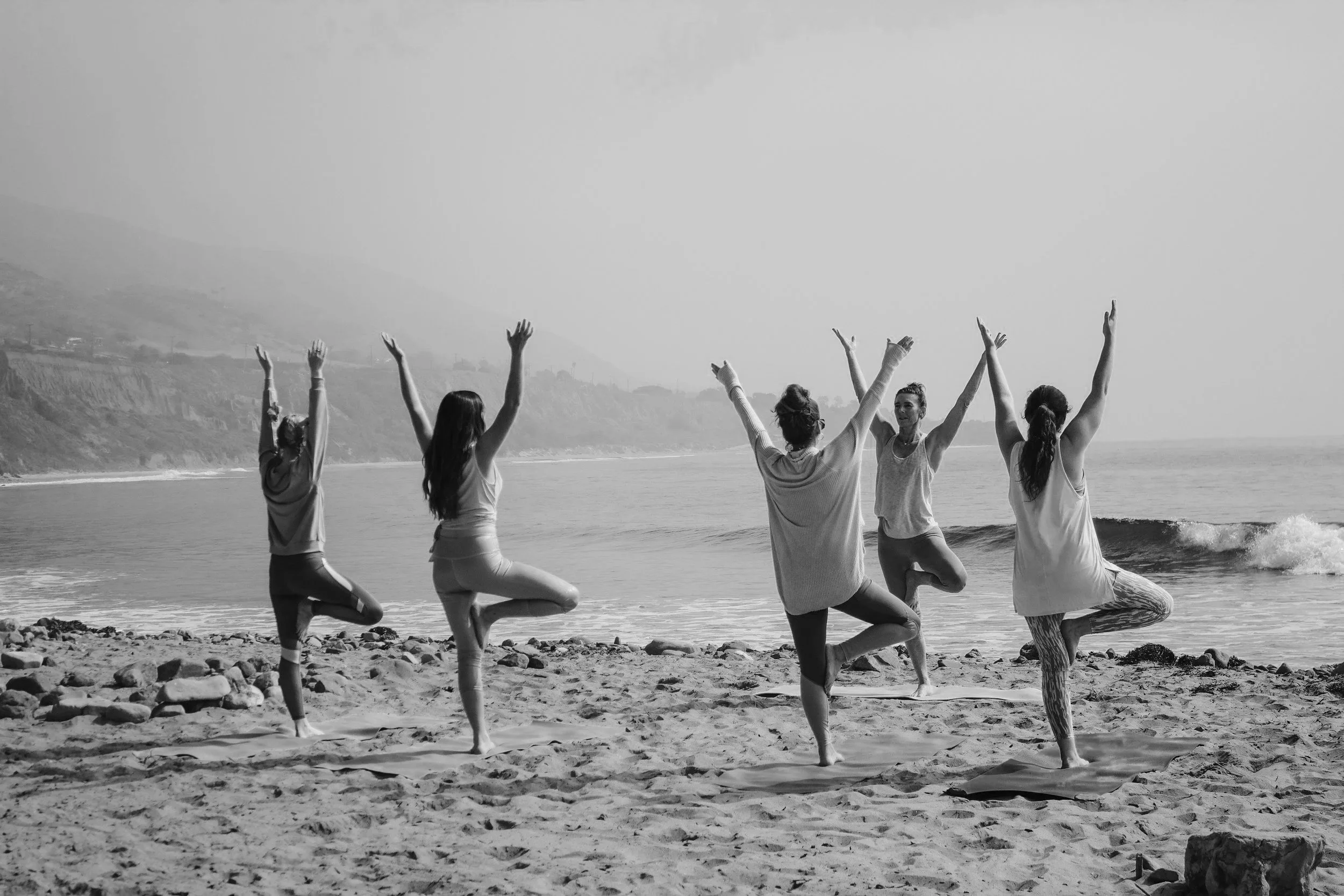 Five women practicing yoga on the beach, standing on yoga mats, facing the ocean, in a tree pose with arms raised overhead.