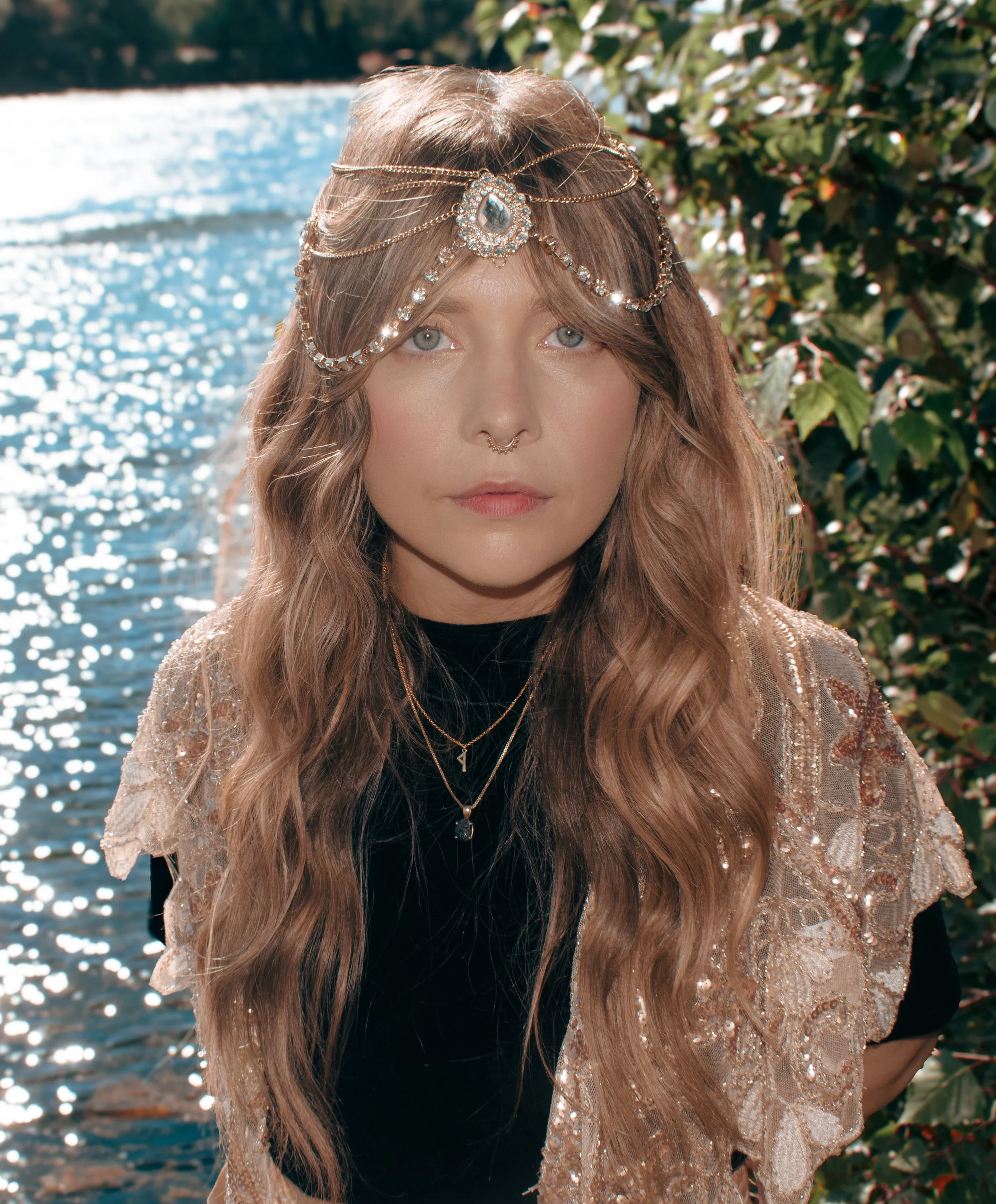 Close-up of a woman with long, wavy brown hair, wearing a decorative headpiece with chains and a large jewel, standing outdoors near water and greenery.