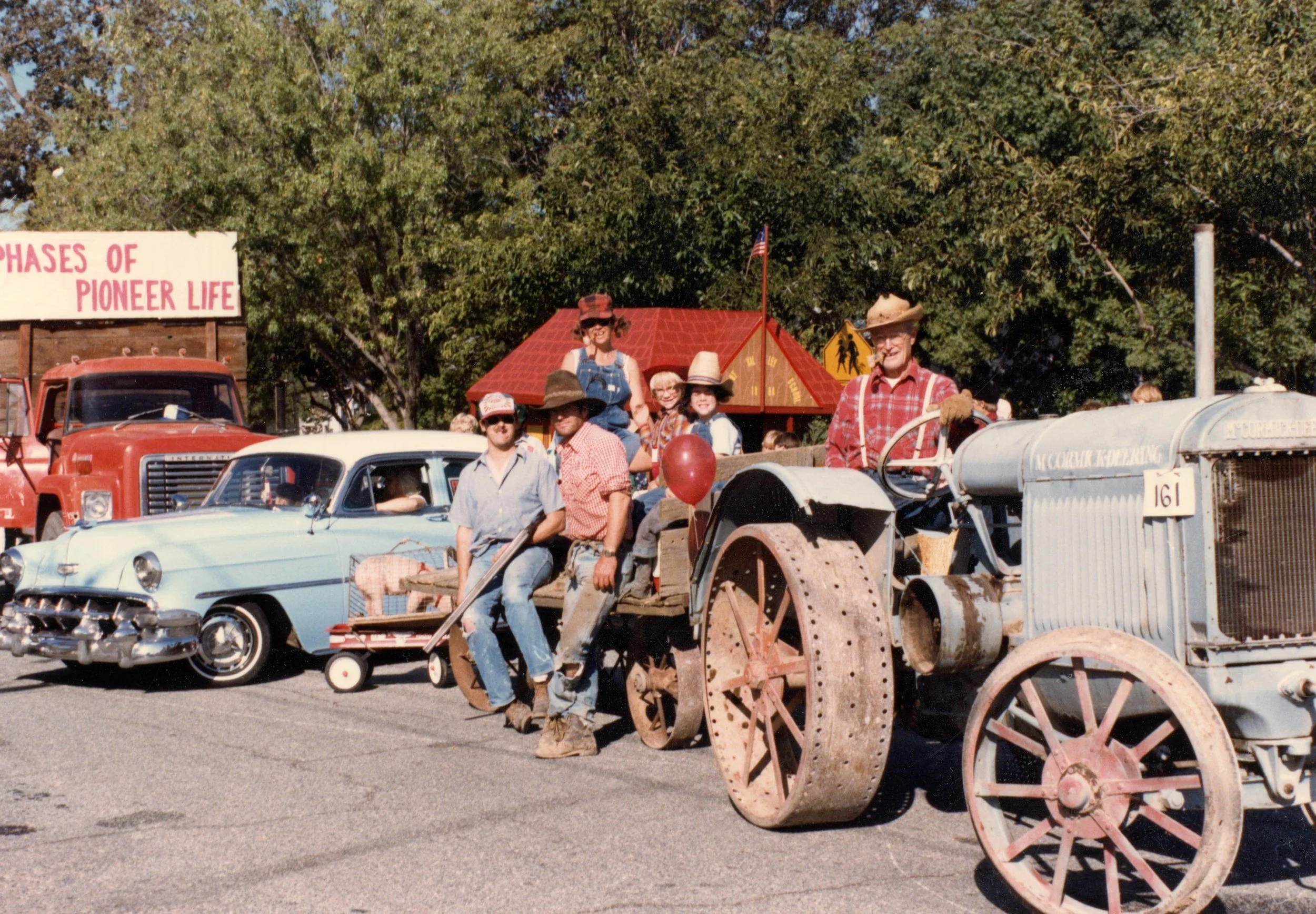 Vintage parade scene with old cars and a tractor, featuring people in cowboy hats and western attire, beside a "Phases of Pioneer Life" float.
