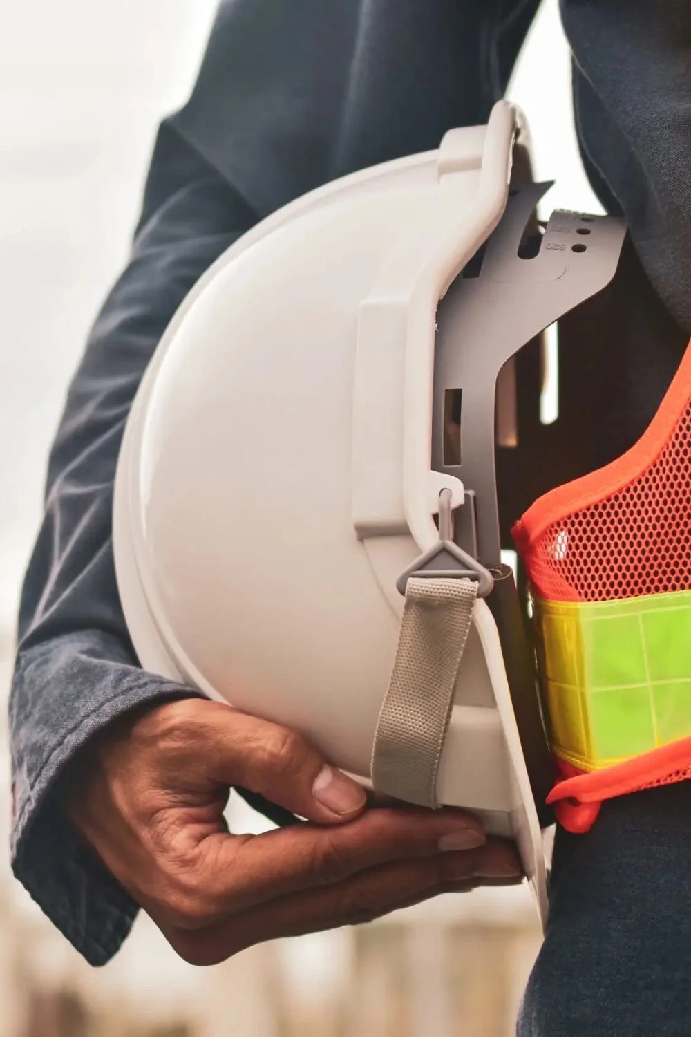 A person wearing a dark jacket and a high-visibility vest holding a white construction helmet.