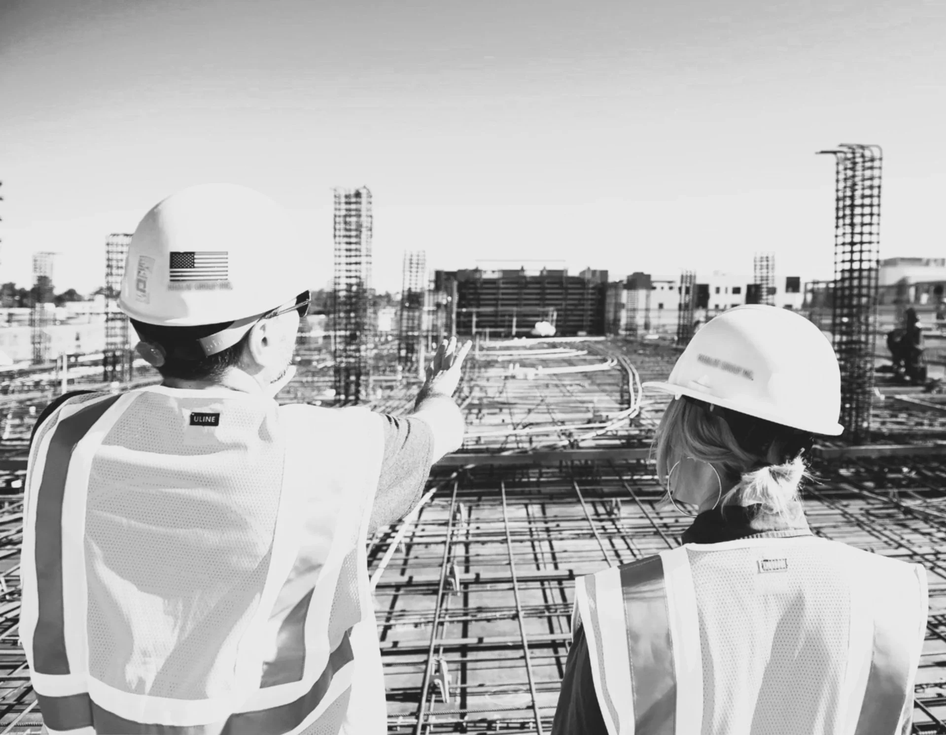 Two construction workers wearing helmets and safety vests inspecting a building under construction on an urban rooftop.