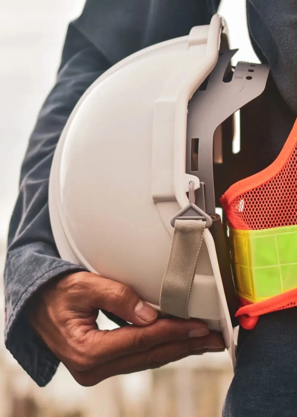 A person holding a white safety helmet with gray straps, wearing a dark gray jacket and a high-visibility safety vest.