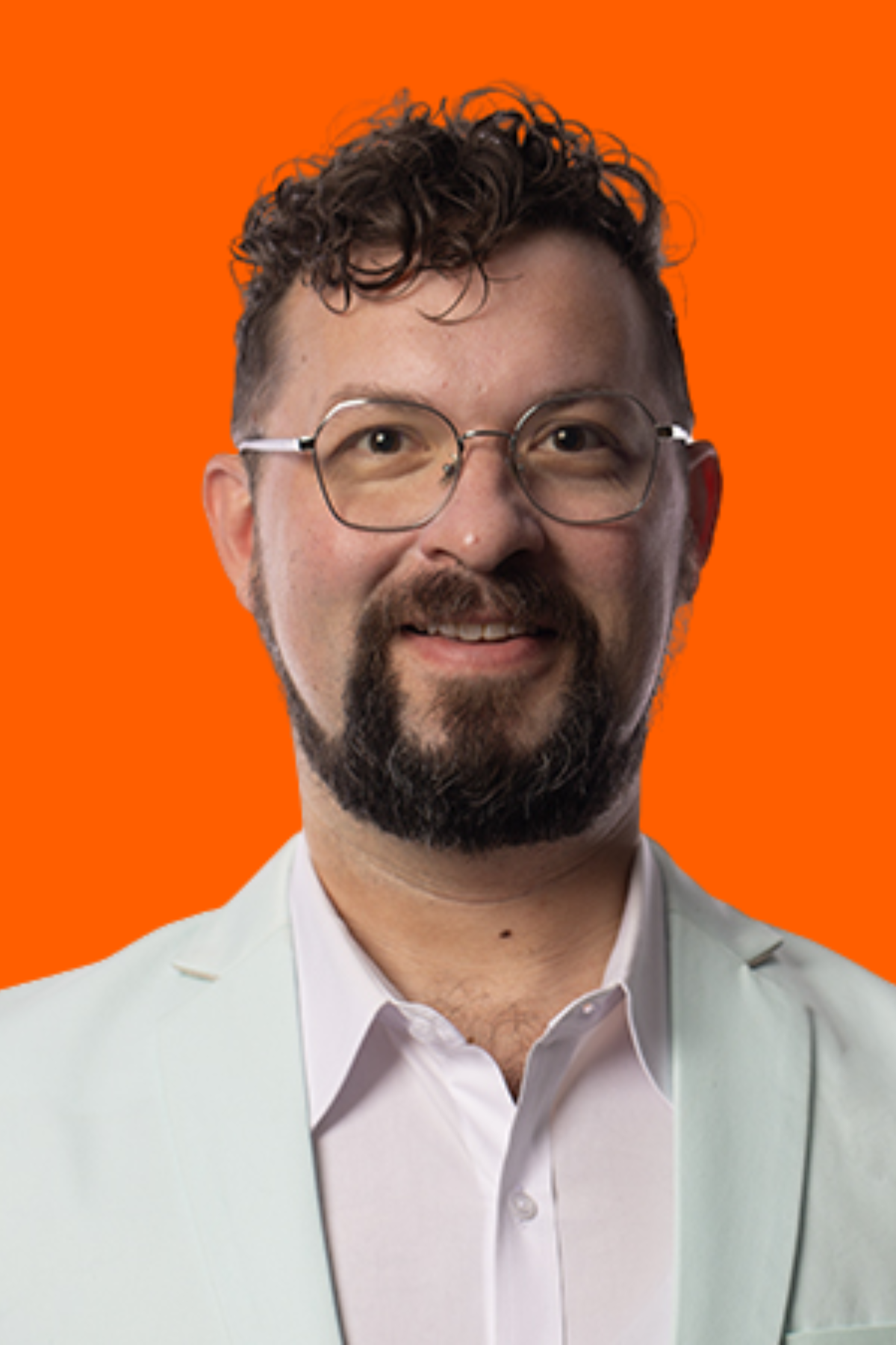 A man with curly hair, glasses, and a beard smiling, wearing a white shirt and cream-colored blazer against an orange background.