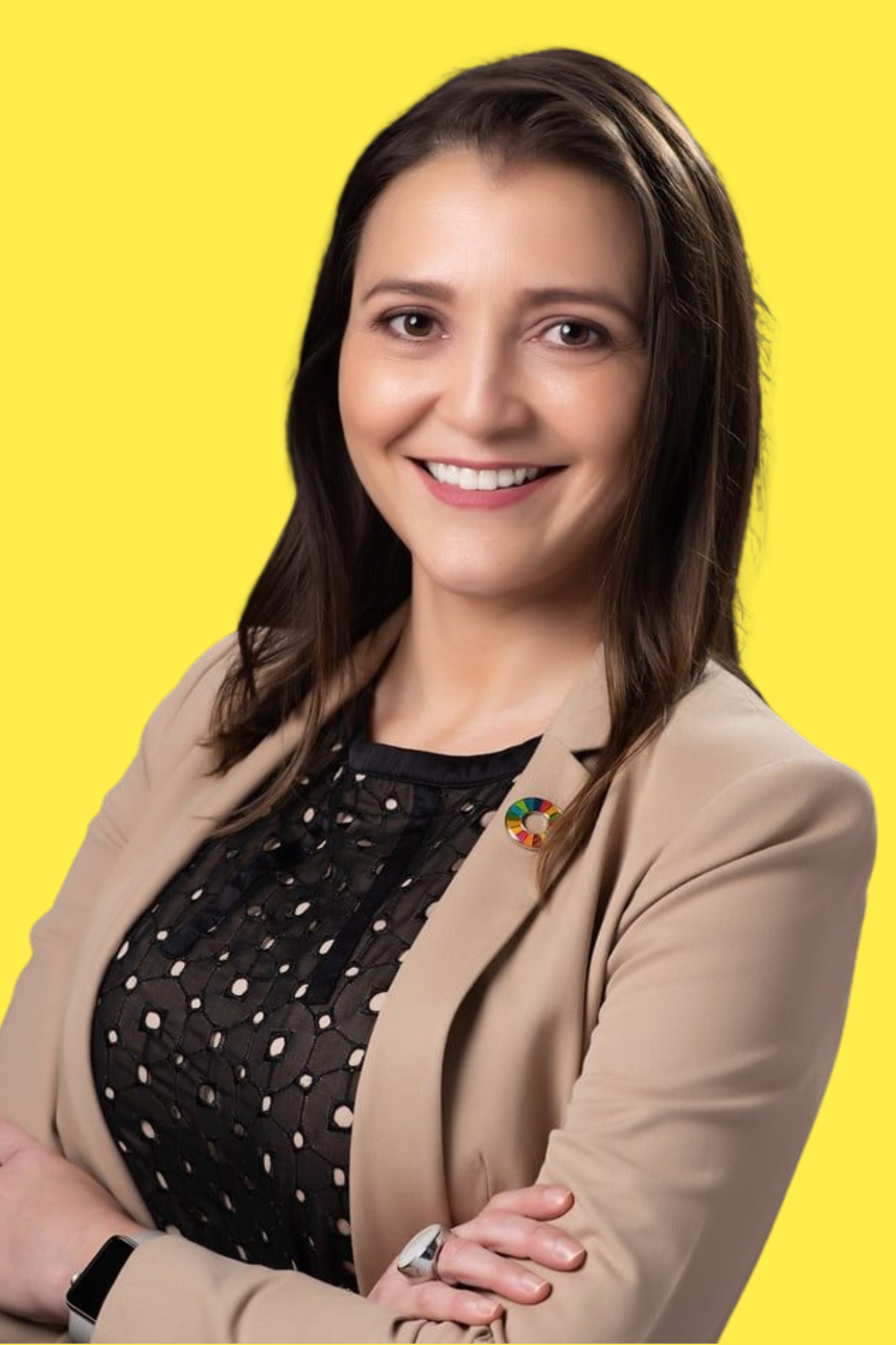 Portrait of a woman with long brown hair smiling, wearing a beige blazer with a colorful circular pin, a black patterned top, standing against a bright yellow background.