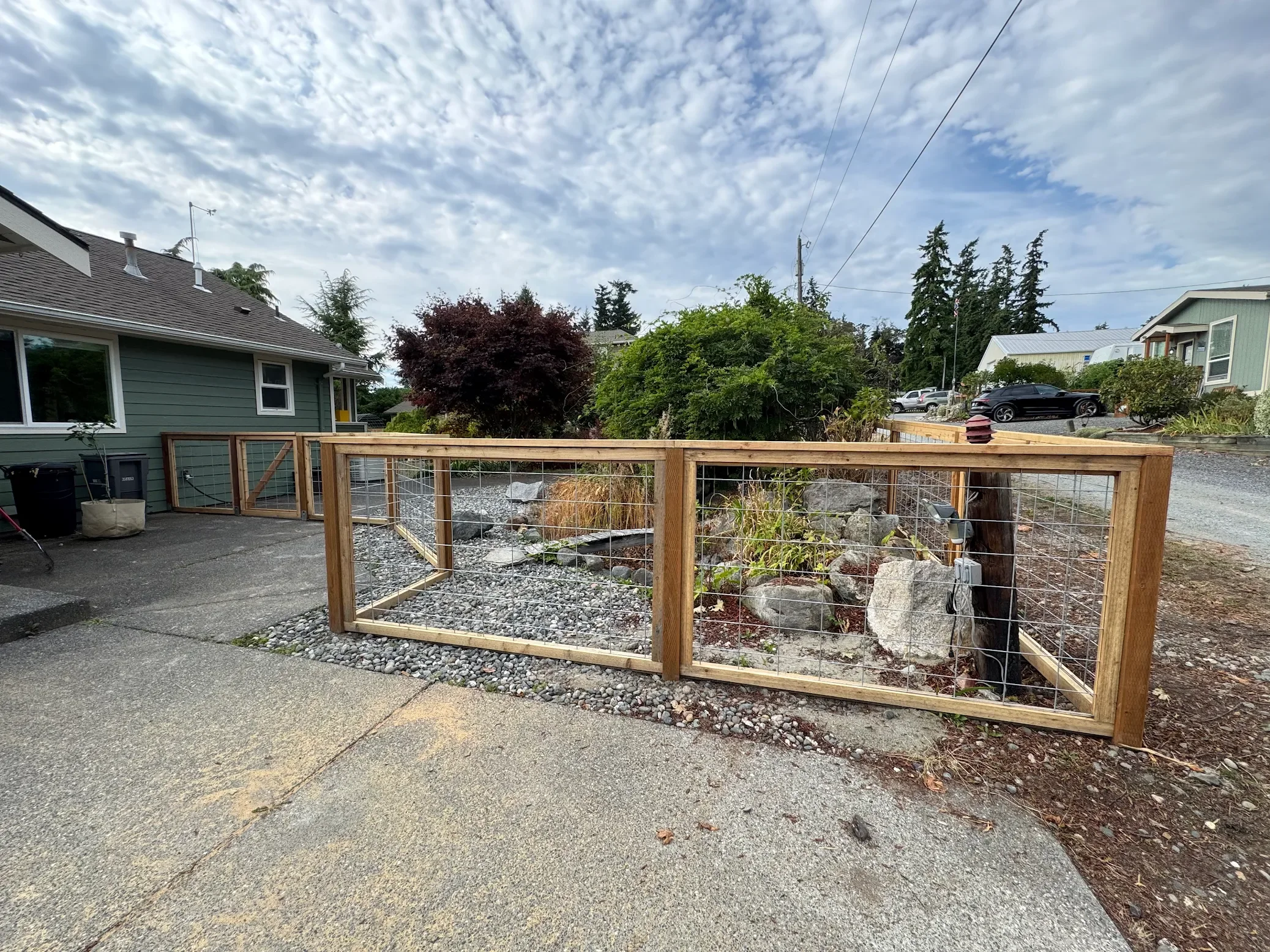 A partially constructed fenced area in a residential driveway, with rocks and plants inside, neighboring houses, trees, and a partly cloudy sky.
