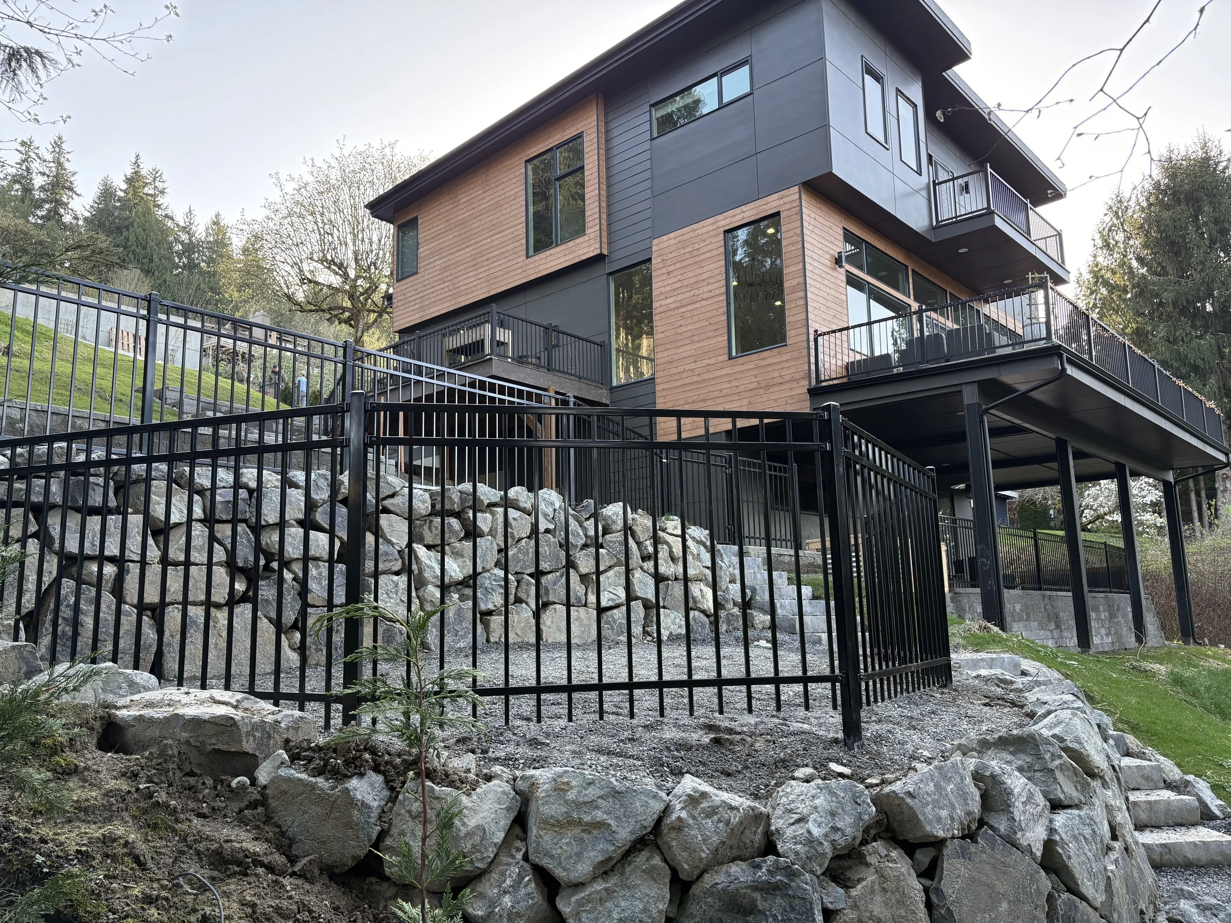 Modern house with black and wood exterior, multiple large windows, and spacious balconies, built on a rocky hillside with a black metal fence and steps leading up to it, surrounded by trees.