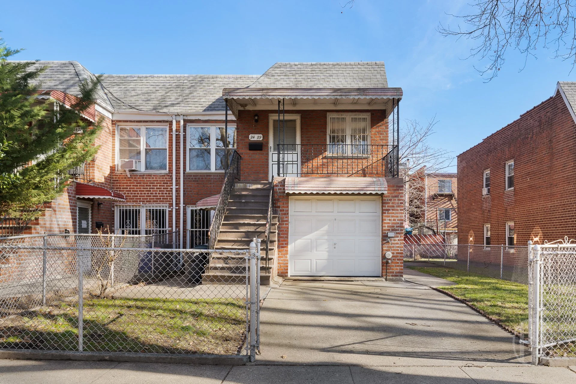 Exterior view of a row of brick apartment buildings with fire escapes, a large tree in front, and parked cars on the street.