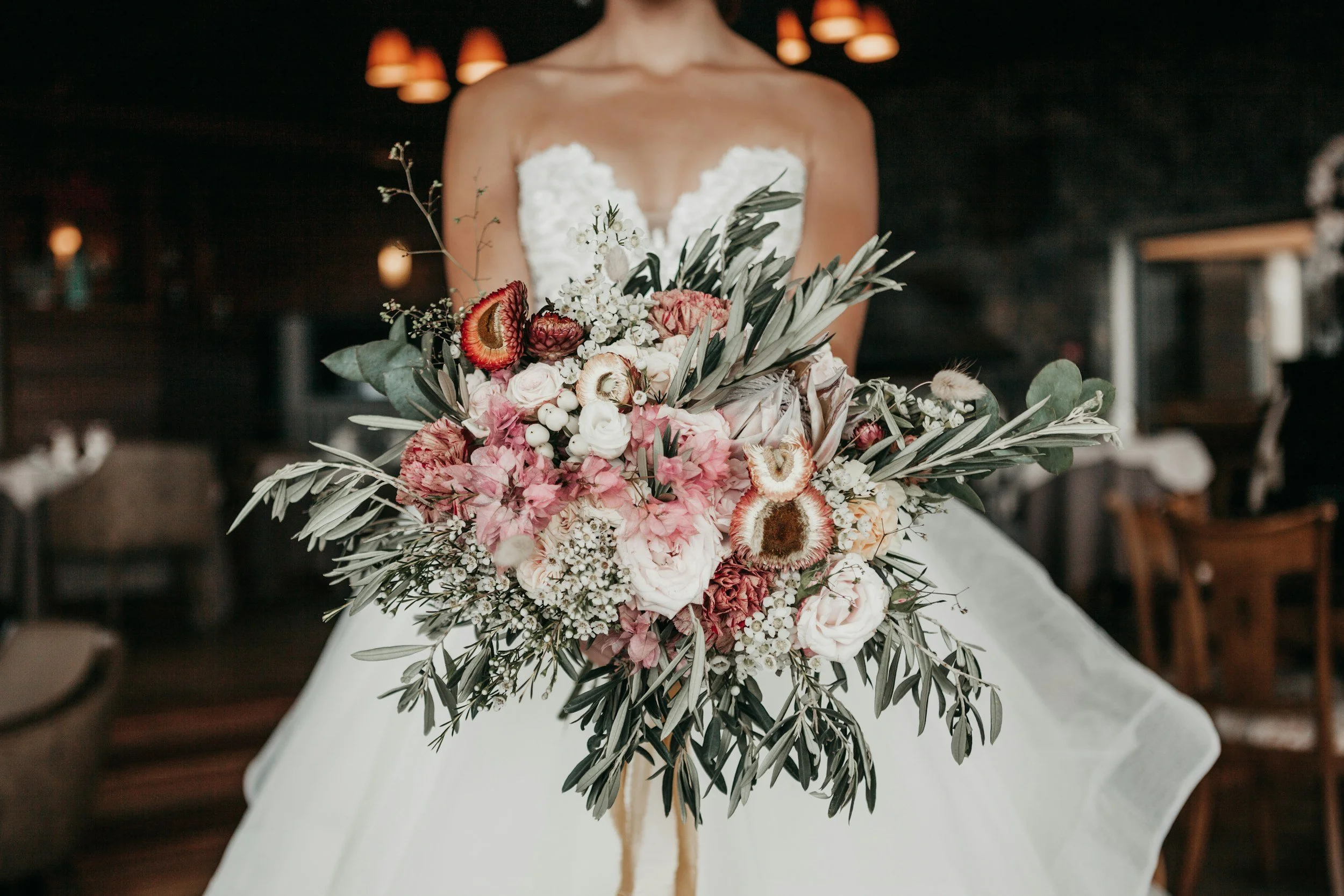 Wedding bride holding large bouquet of pink, white, and red flowers with green foliage, in a room with dim lighting and wooden furniture.