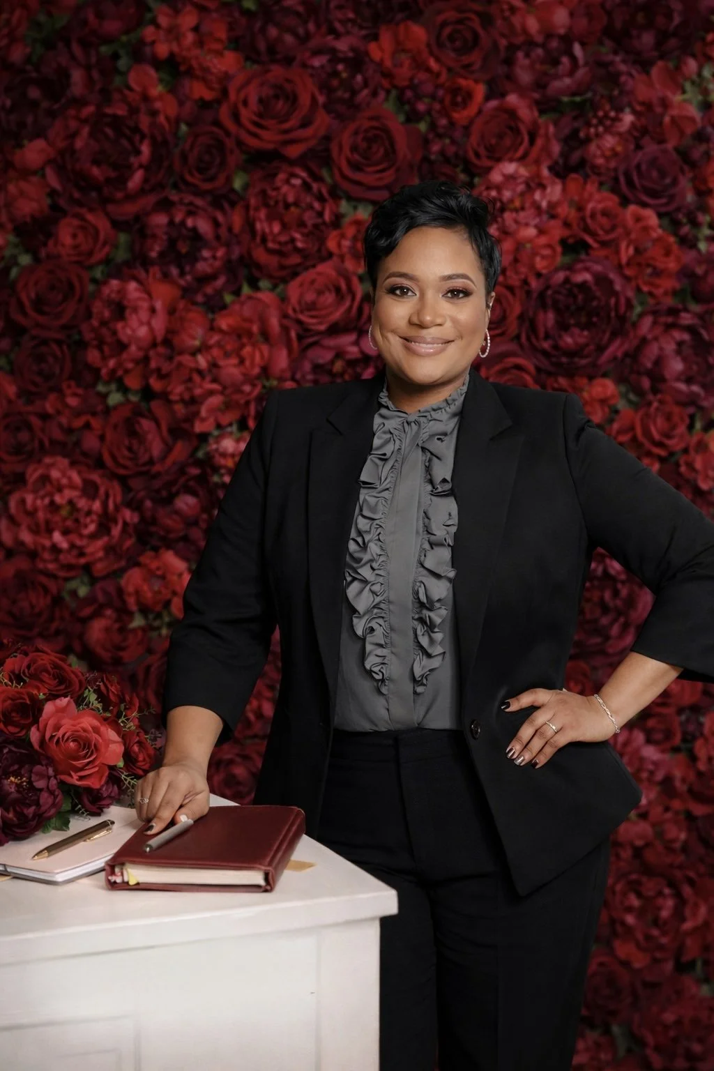 A woman smiling and posing with her hand on her hip, standing next to a white table with a notebook, pen, and flowers, in front of a background wall covered with red roses and dark red peonies.