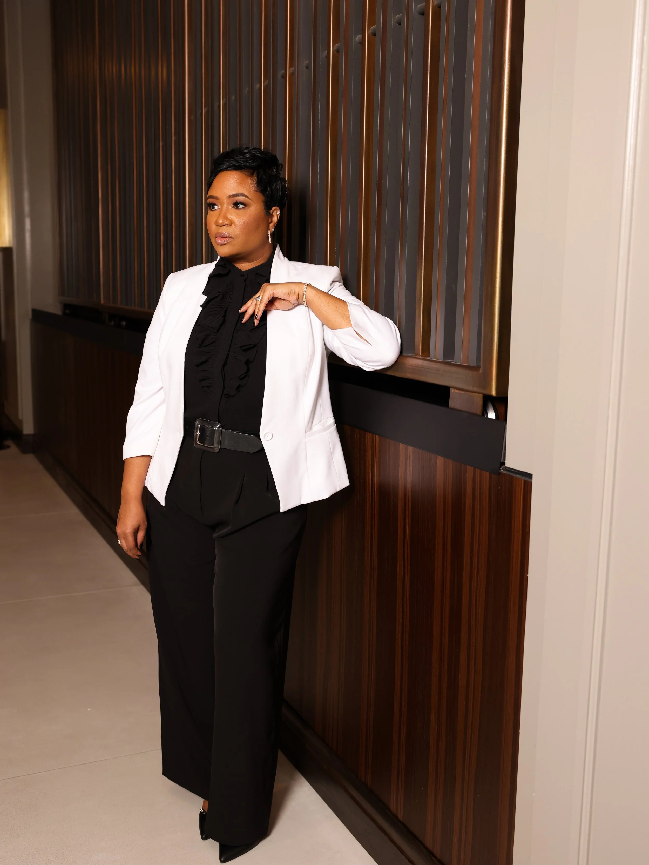 A woman in a formal black and white outfit stands leaning against a wooden paneled wall.