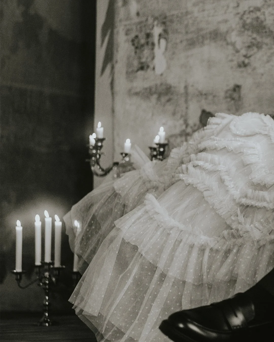 Ein schwarz-weiß foto zeigt eine alte Wand mit einer Kerzenleuchter mit brennenden Kerzen und einen Tisch mit einem weißen Tüllkleid, das an einer Person getragen wird, deren Schuhwerk sichtbar ist.
